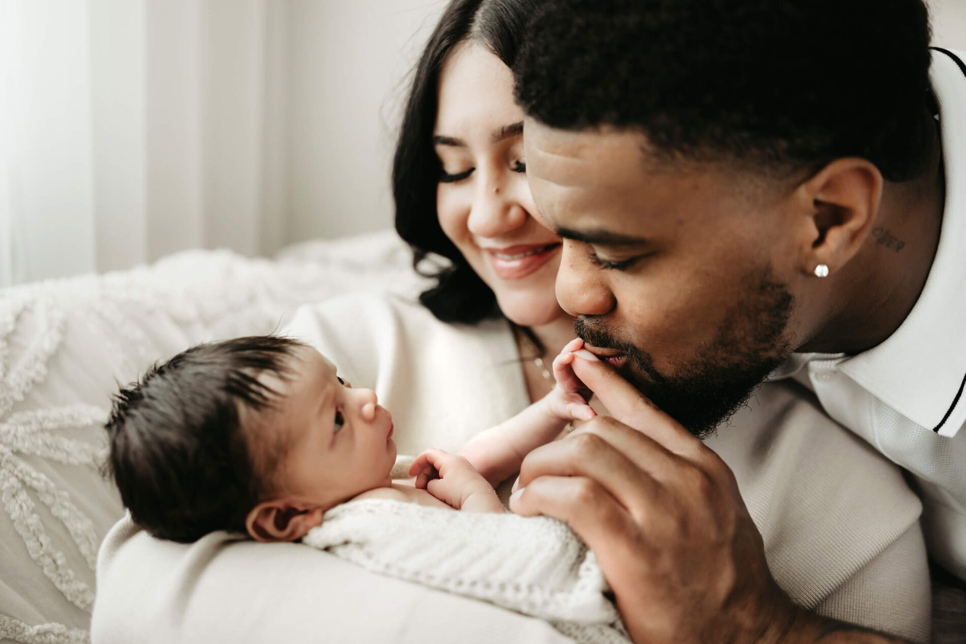 One parent kisses an awake newborn’s toes while the other smiles beside them on a white bed.
