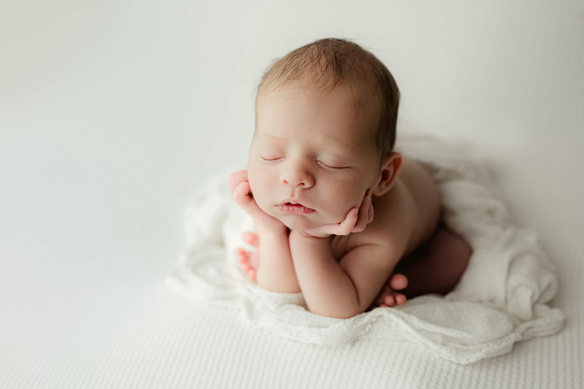 Unwrapped newborn with eyes closed rests with chin in both hands on a white textured cushion.