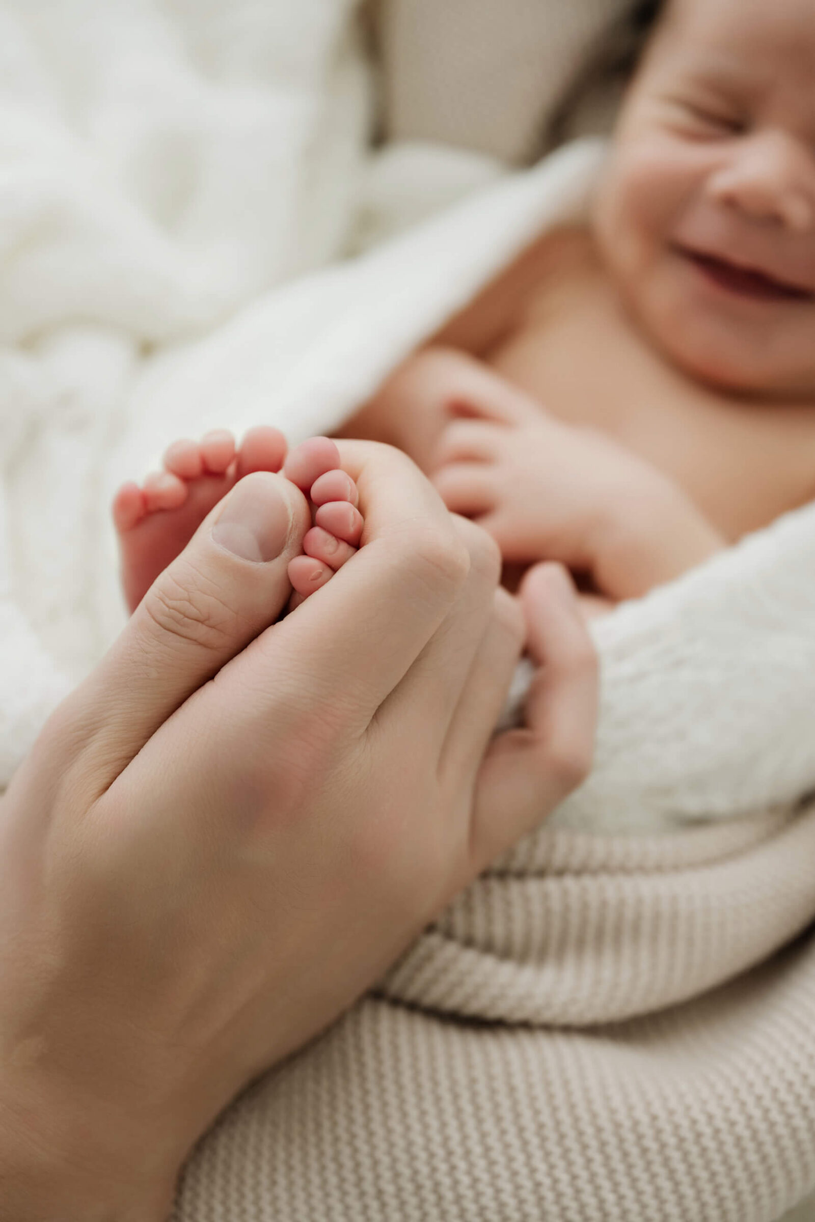 Adult hand holds a newborn’s tiny toes, with the baby’s face softly blurred in the background.
