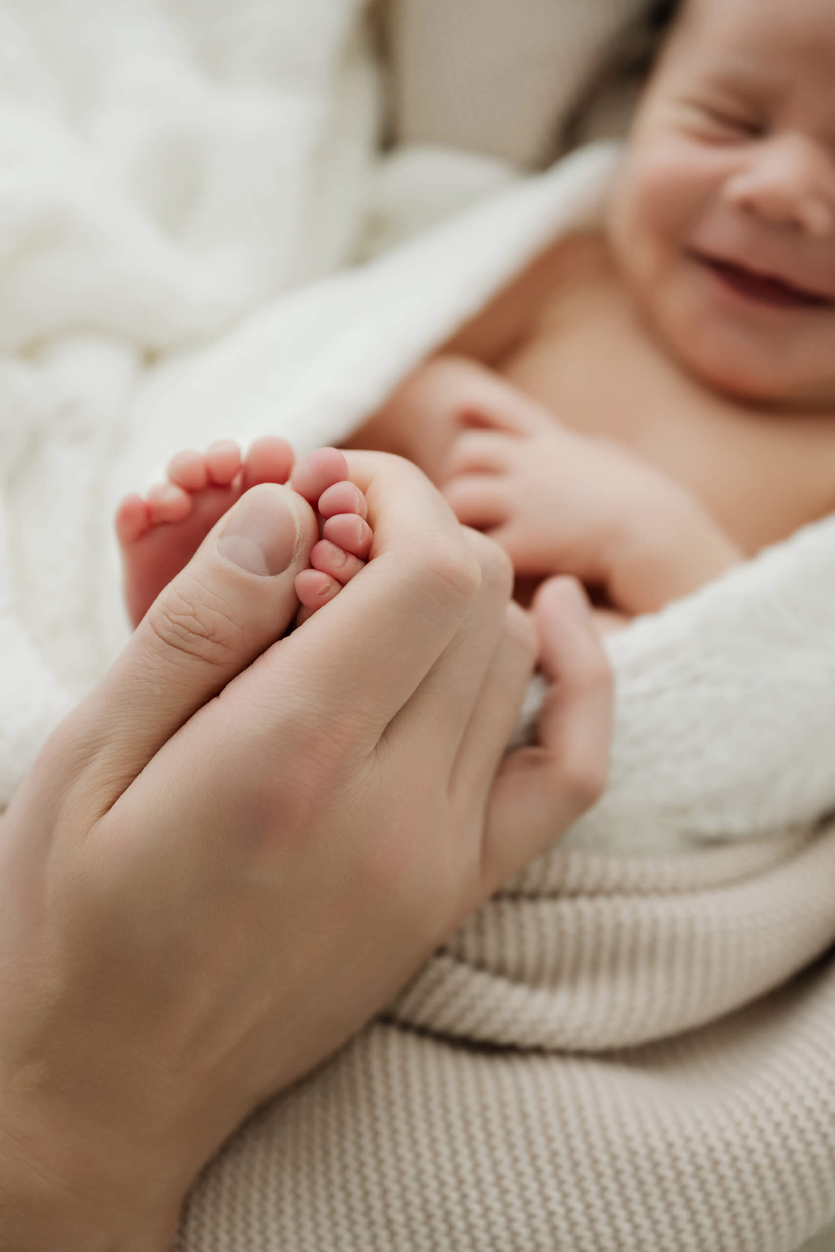 Adult hand holds a newborn’s tiny toes, with the baby’s face softly blurred in the background.