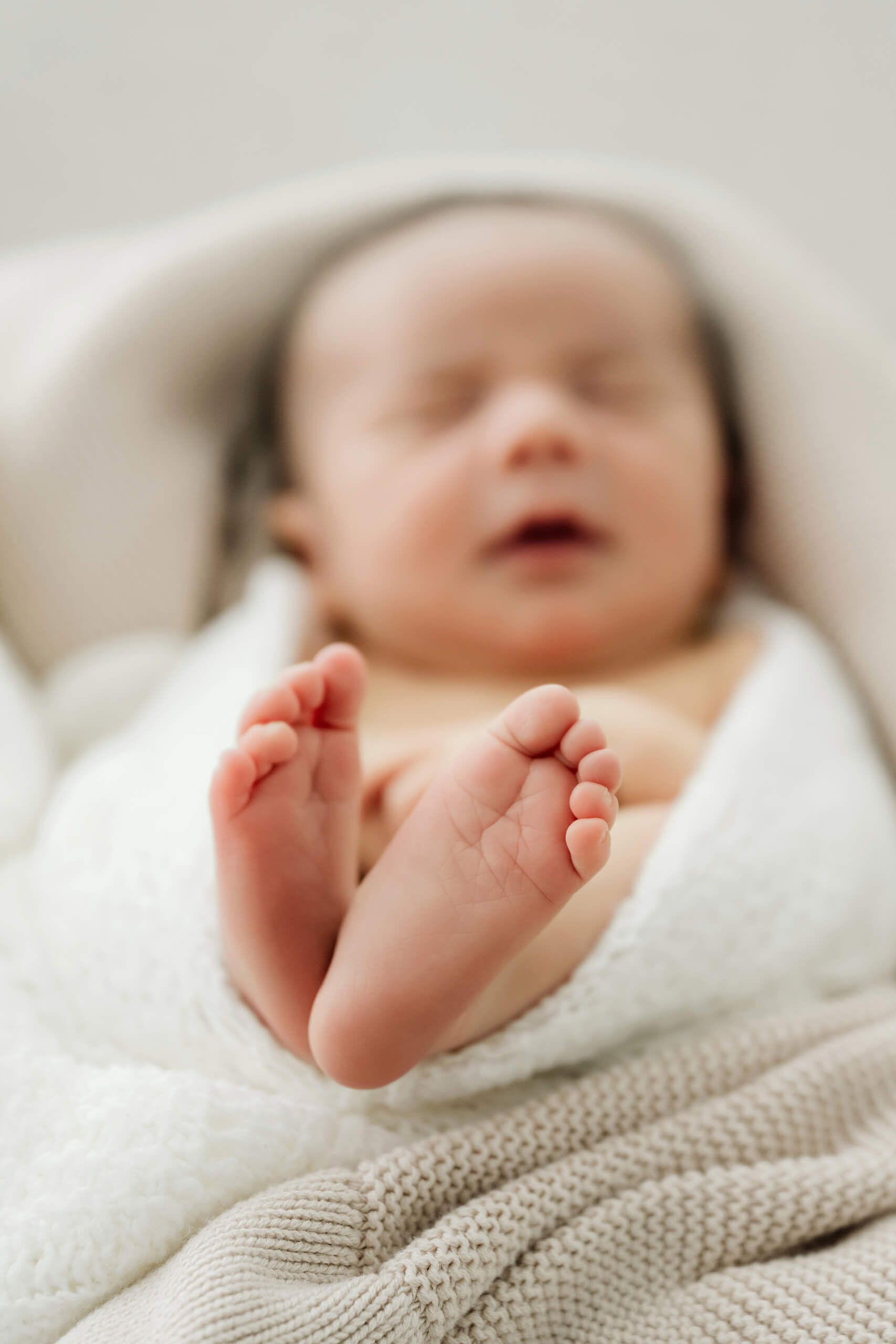 Newborn’s bare feet fill the foreground while the baby’s face remains softly blurred behind.