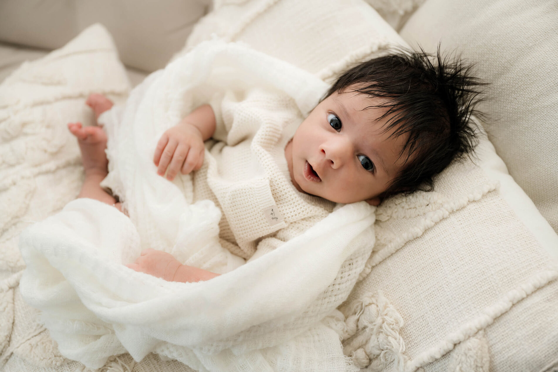Awake newborn in a cream knit outfit lies among white pillows and blankets, looking toward the camera.
