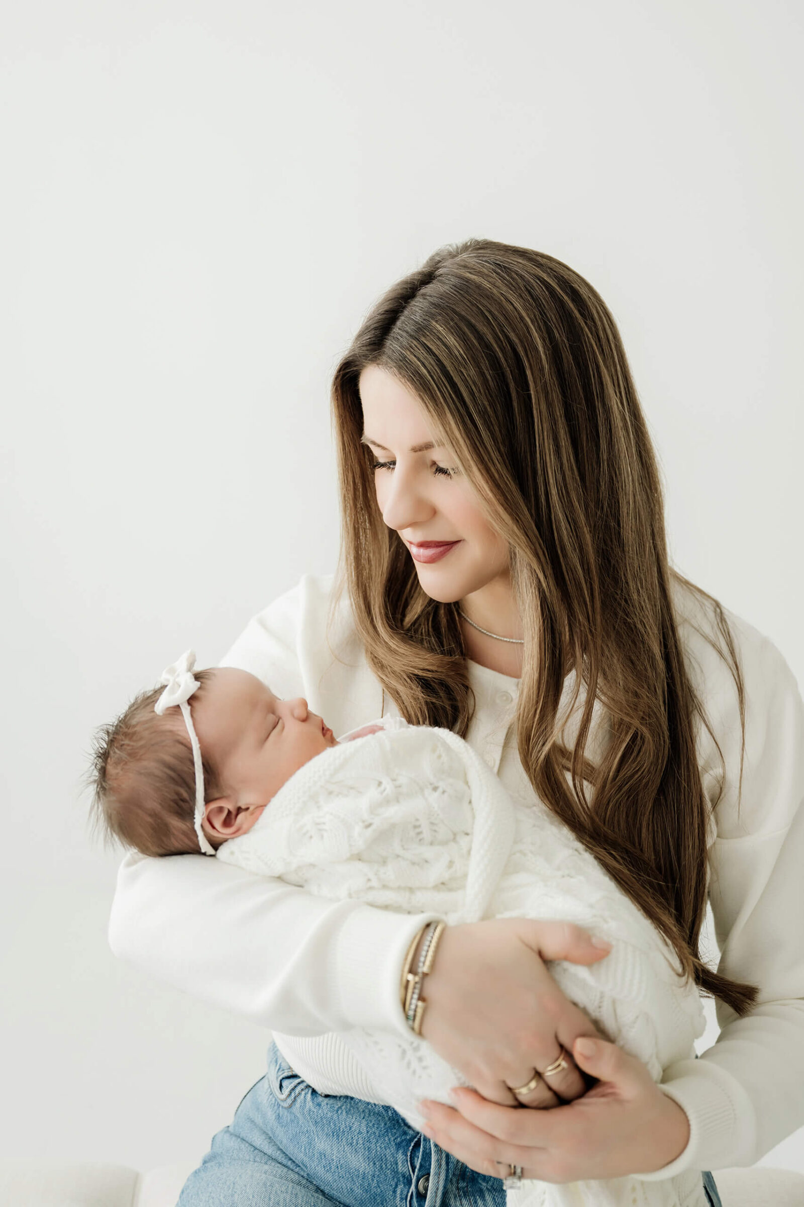 Parent looks down while cradling a newborn in a white knit blanket and bow headband.