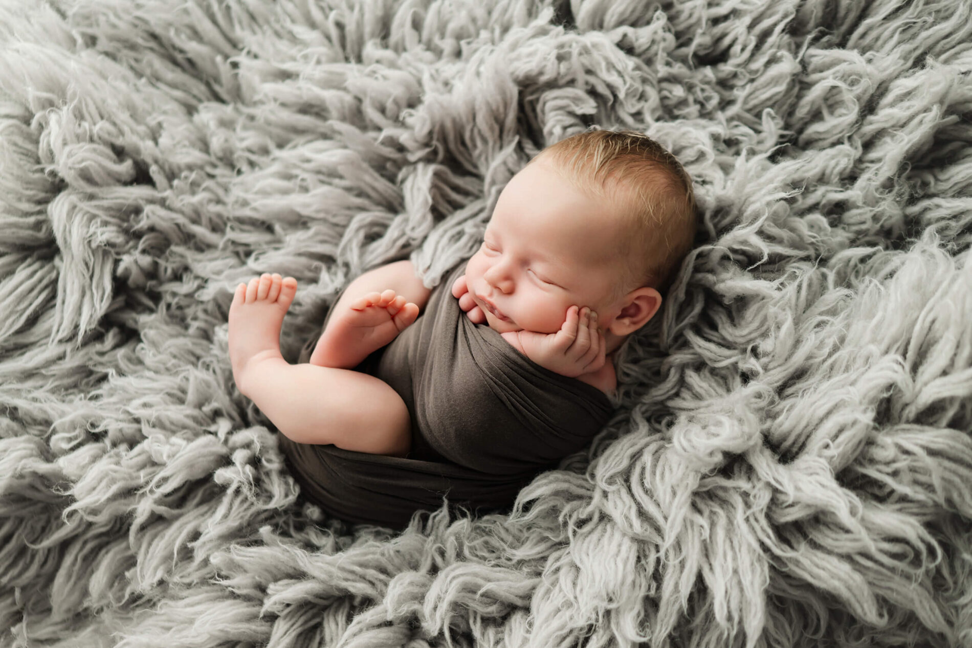 Newborn with eyes closed rests in a dark gray wrap on a shaggy gray blanket.