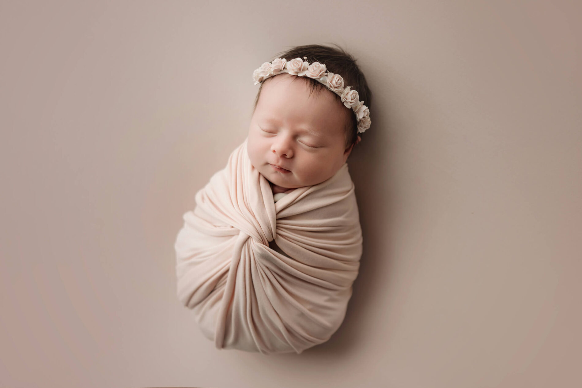 Pale pink-wrapped newborn with eyes closed wears a small floral headband on a blush backdrop.