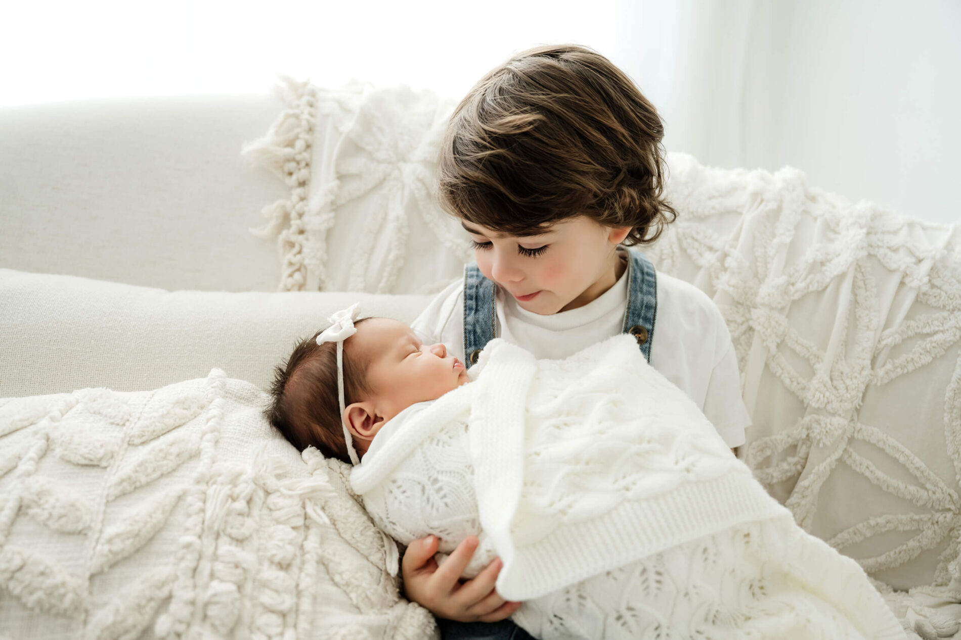 Young sibling looks down while holding a newborn bundled in a white knit blanket.
