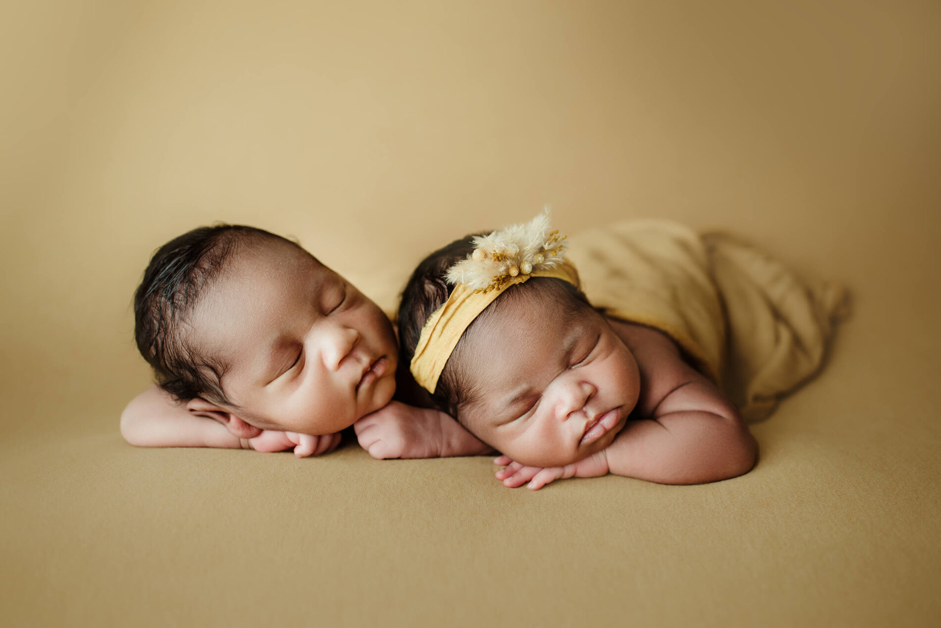 Two newborns with eyes closed rest side by side on a mustard-colored backdrop.