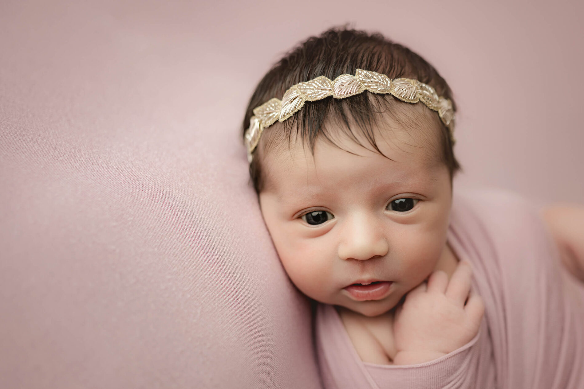 Awake newborn wrapped in dusty pink wears a gold leaf headband in a close portrait.