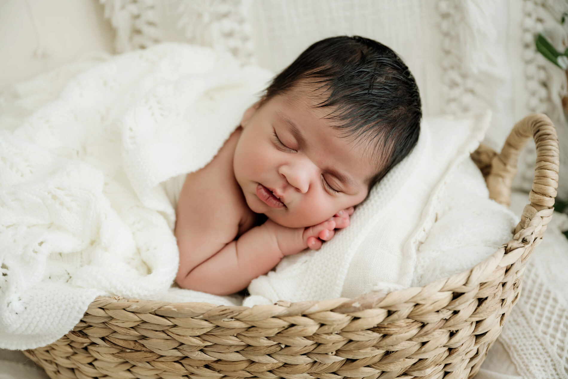 Newborn with eyes closed rests among white knit blankets inside a woven basket.