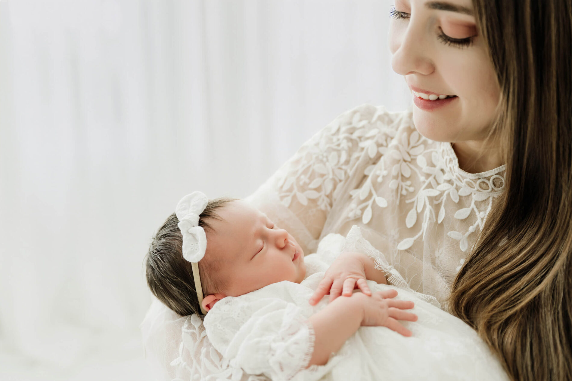 Parent smiles down at a newborn in a white lace outfit and bow headband.