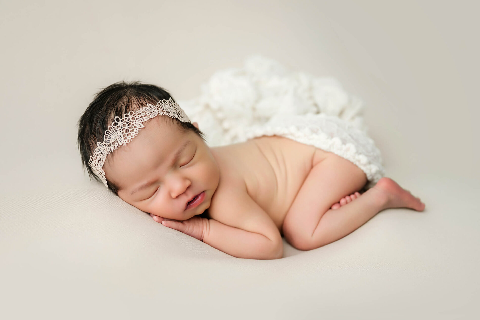 Unwrapped newborn with eyes closed rests on folded arms, wearing a white lace headband and textured bloomers.