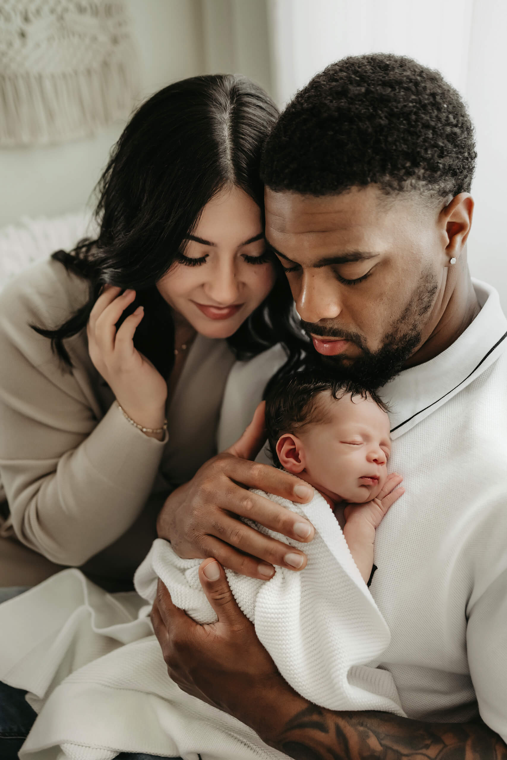 Two parents look down at their newborn resting against one parent’s chest in a white knit blanket.