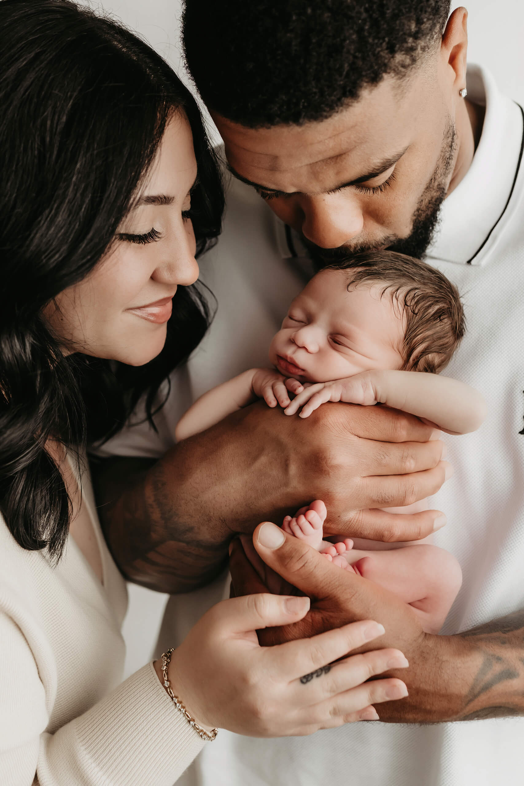 Two parents cradle an unwrapped newborn while one kisses the baby’s head against a light background.