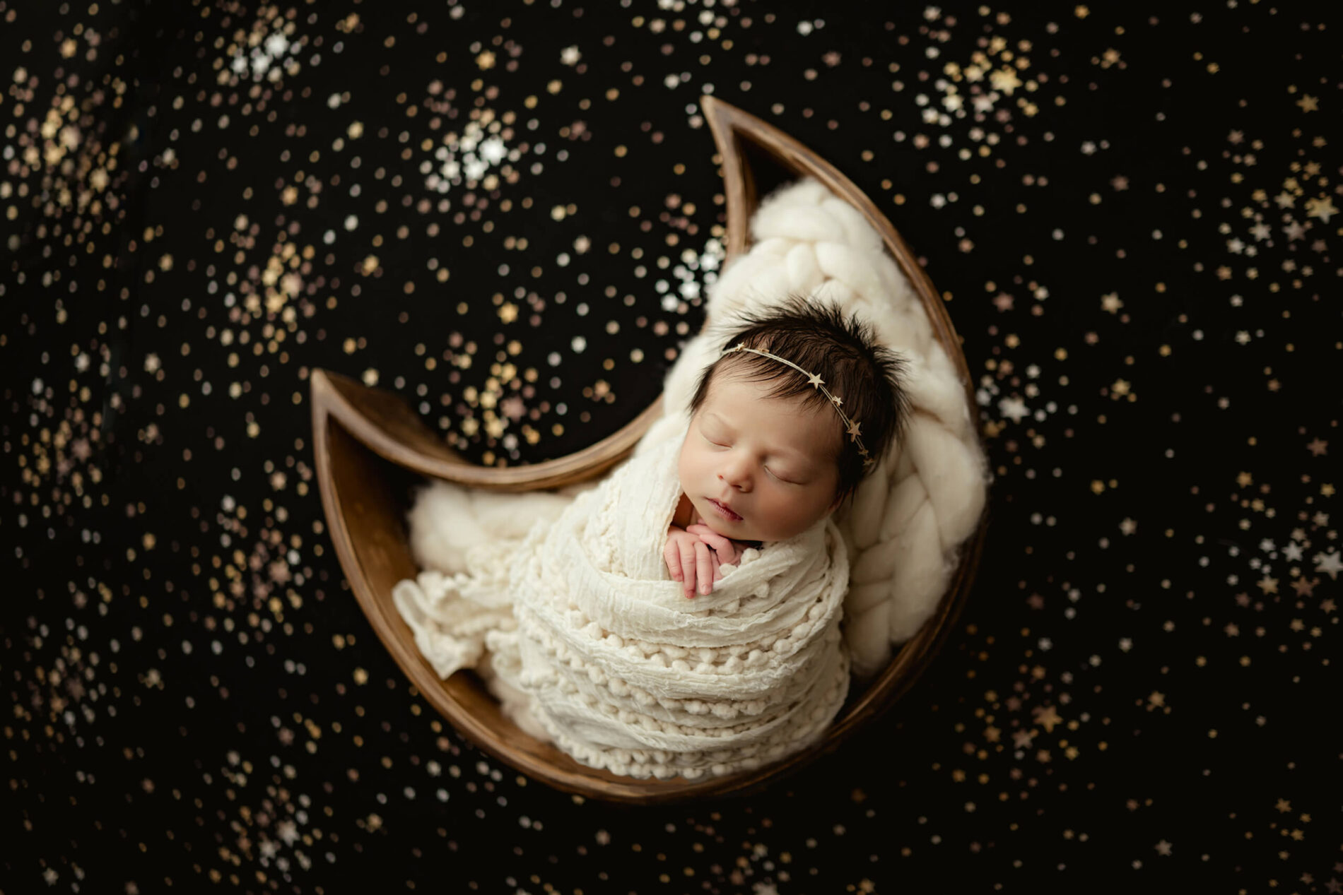 Cream-wrapped newborn with eyes closed rests in a crescent moon prop on a star-patterned black backdrop.