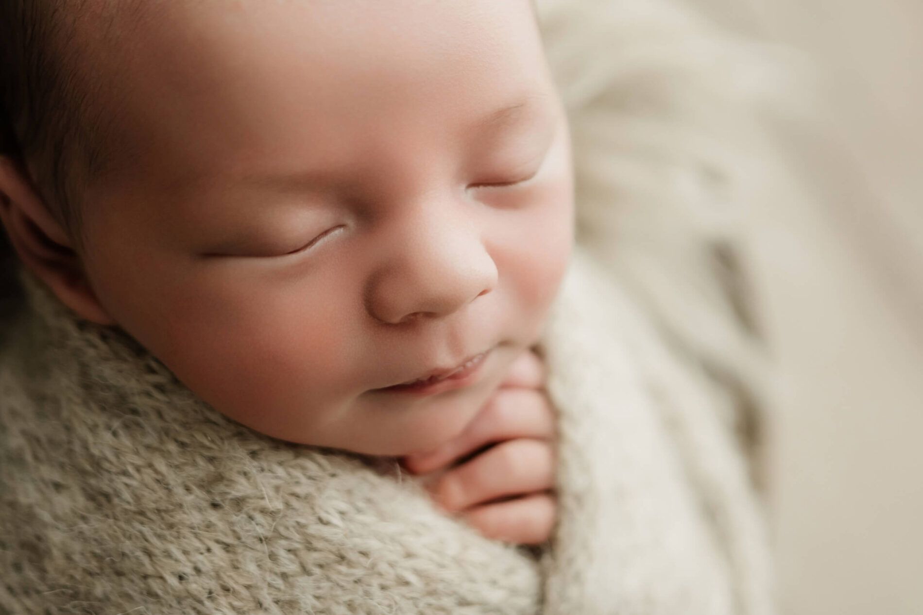 Close-up of a newborn with eyes closed, chin resting on folded hands against a beige knit blanket.