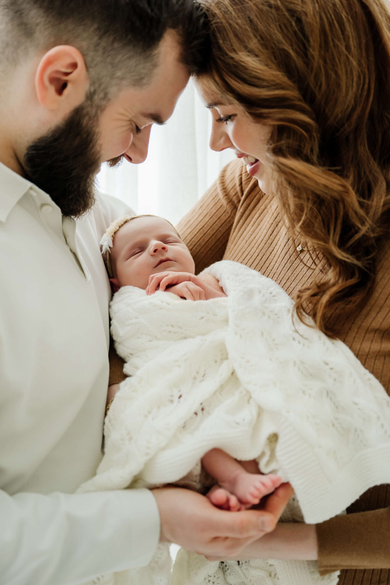Two parents smile down at their newborn wrapped in a white knit blanket, with tiny feet visible.