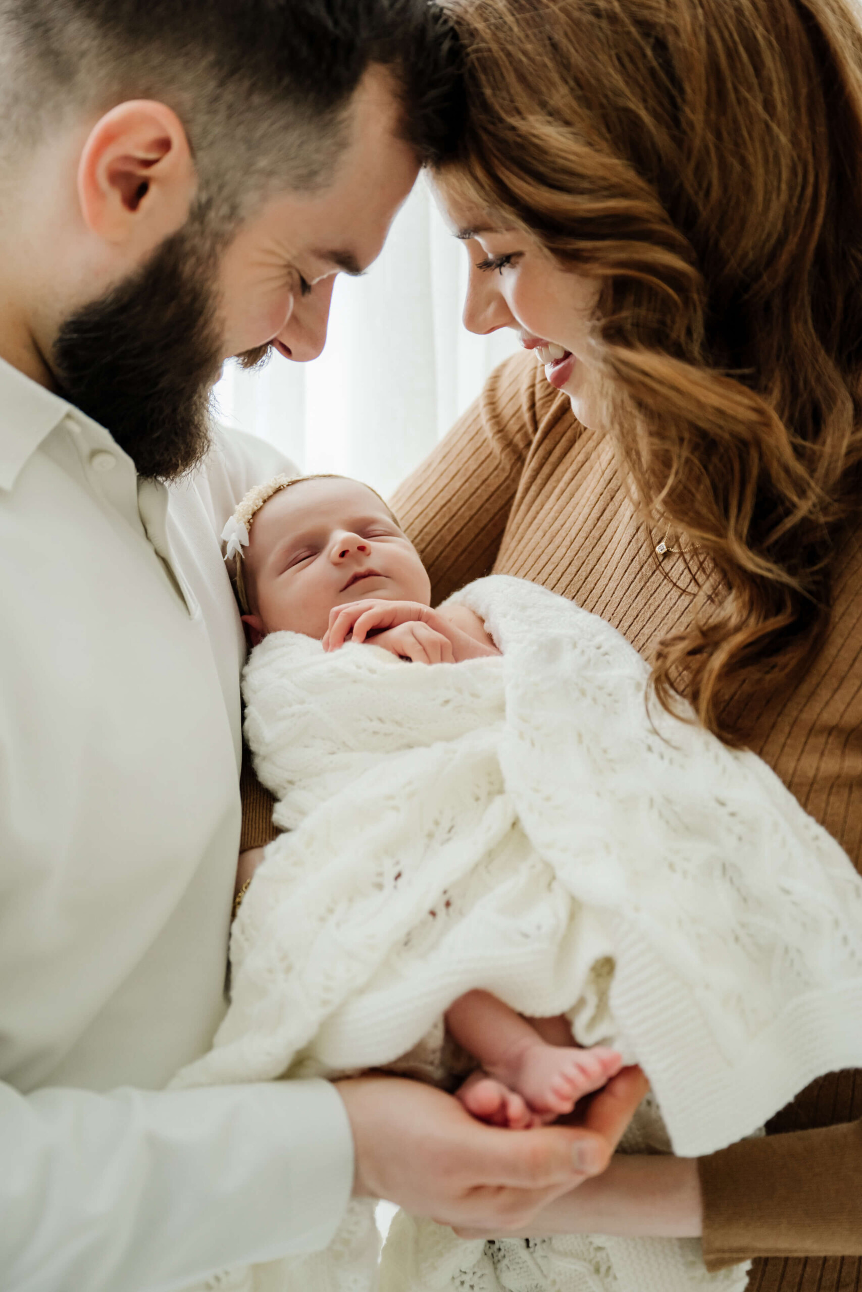 Two parents smile down at their newborn wrapped in a white knit blanket, with tiny feet visible.