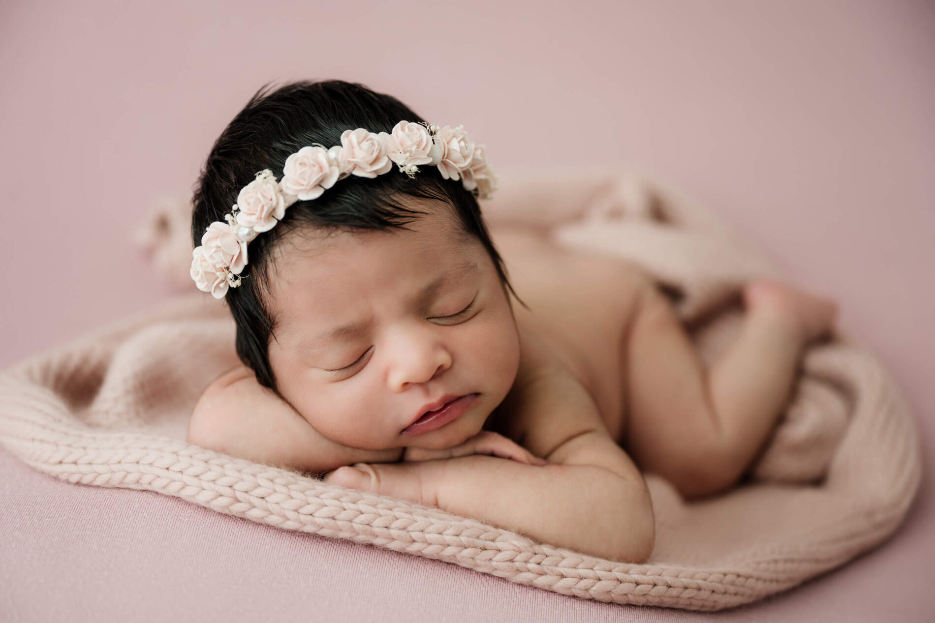 Unwrapped newborn with eyes closed rests on folded arms, wearing a pale floral headband on a pink backdrop.