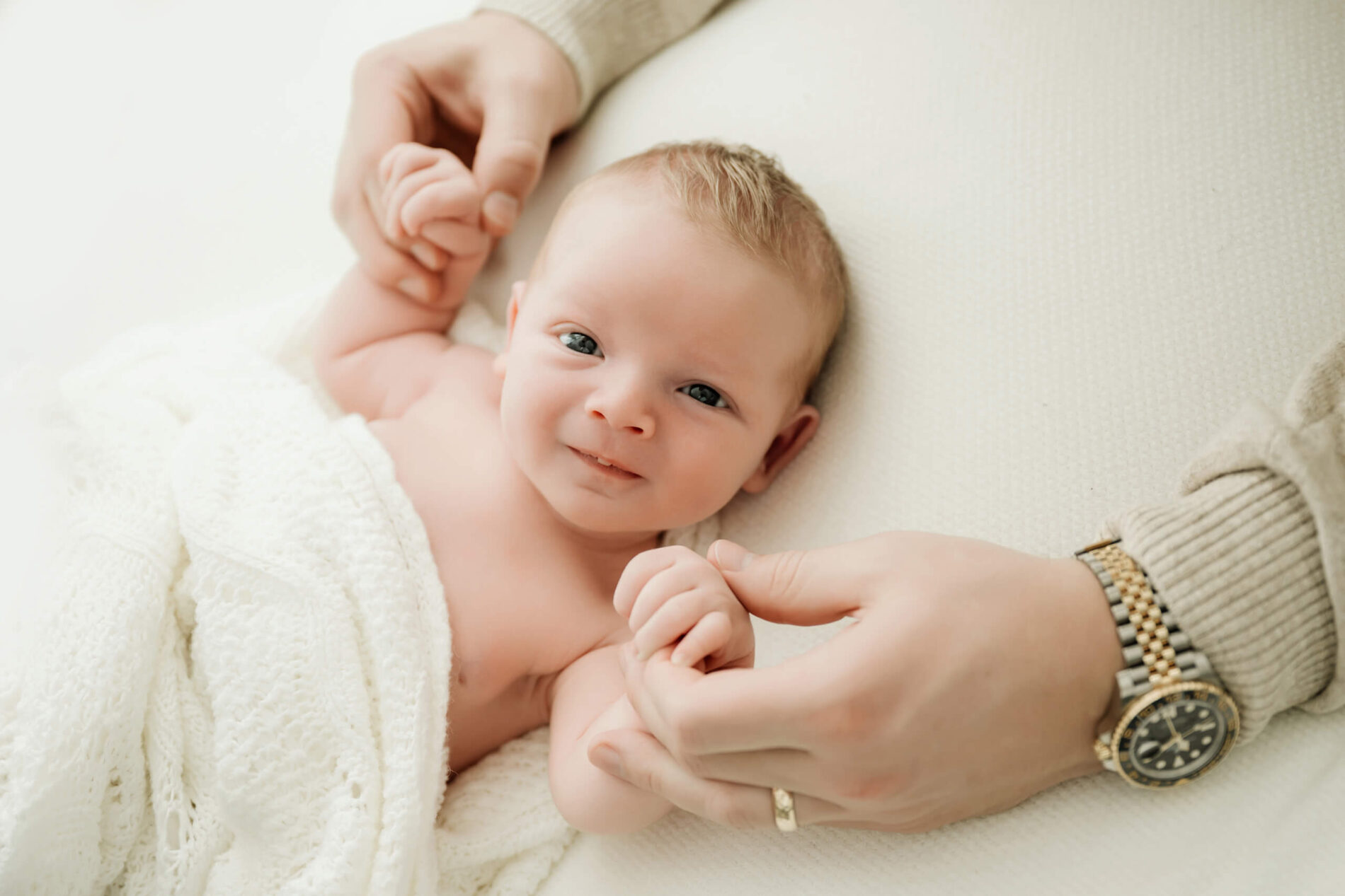 Awake newborn lies beneath a white blanket while two adults gently hold the baby’s hands.