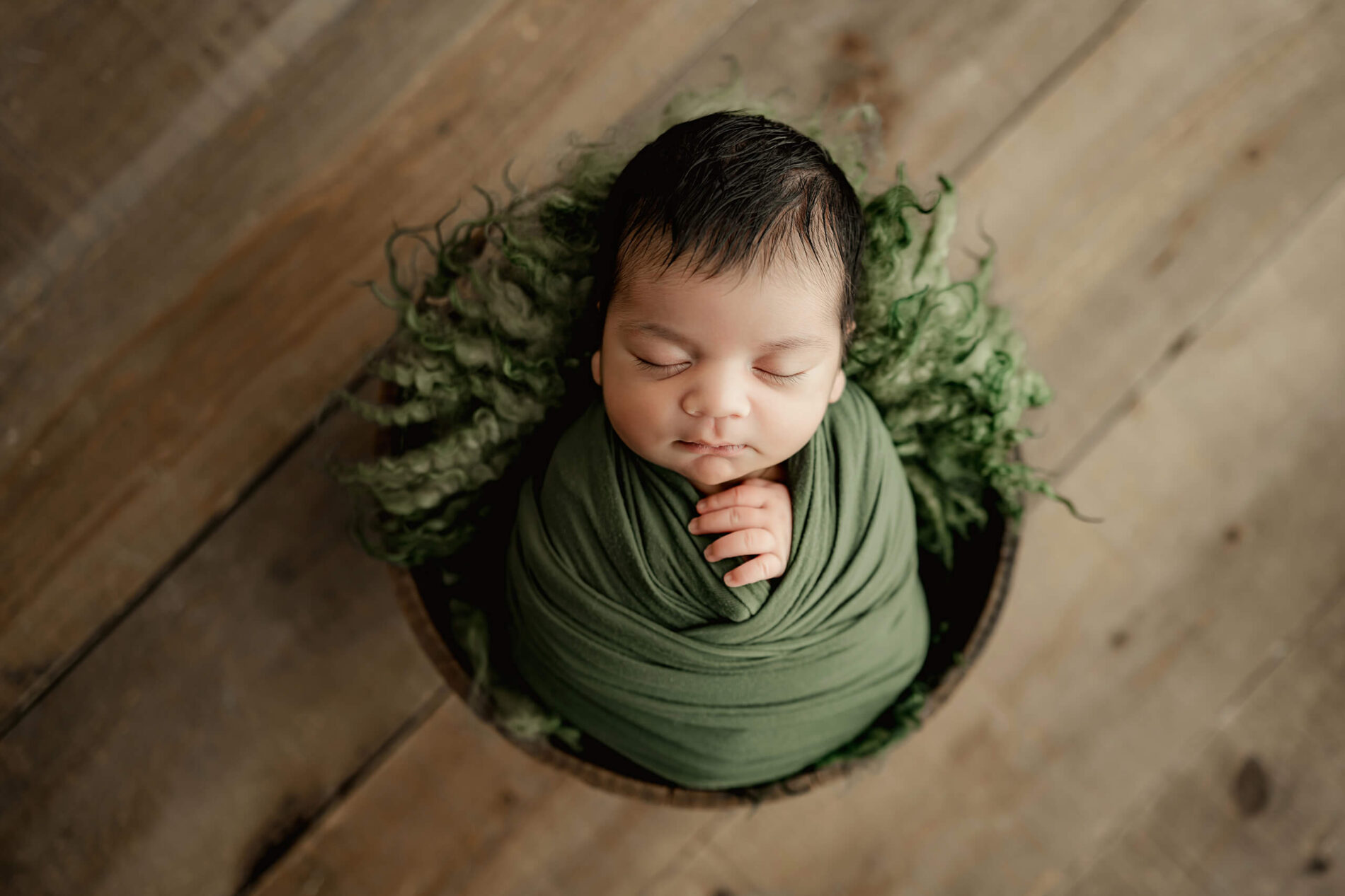 Green-wrapped newborn with eyes closed rests in a round bowl surrounded by textured green fibers.