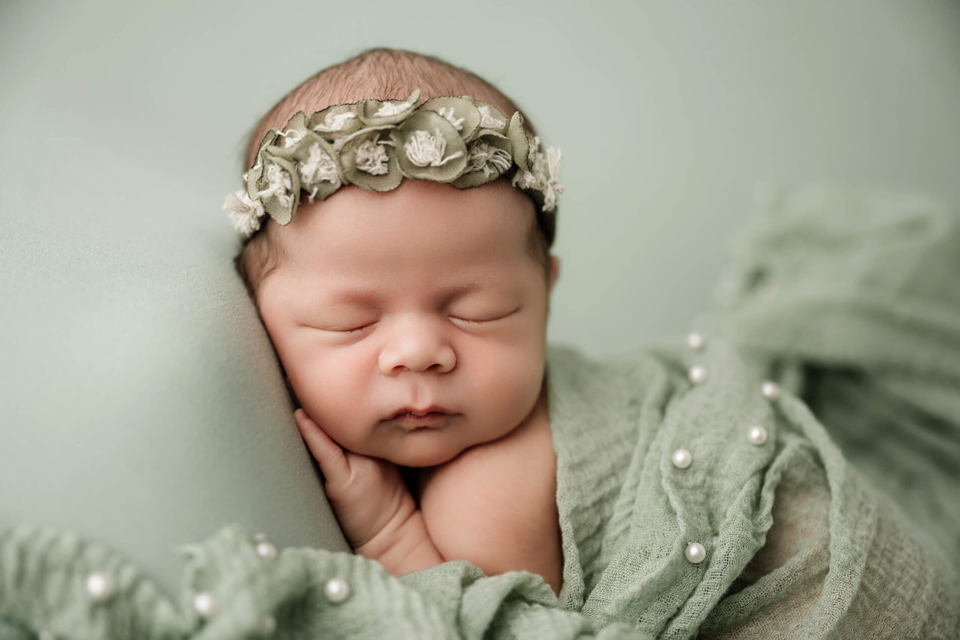 Newborn with eyes closed wears a green floral headband beside sage fabric accented with small pearls.
