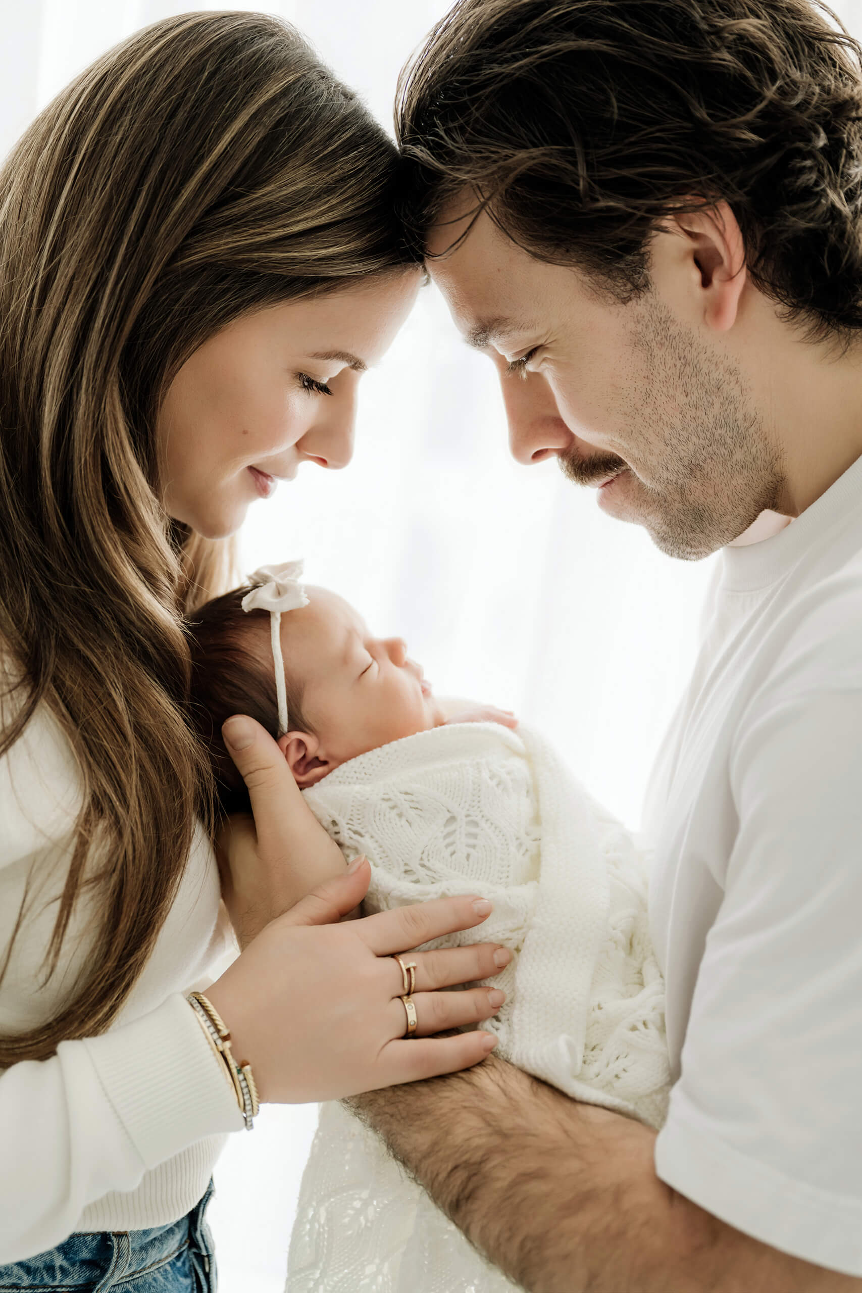 Two parents lean close and look down at their newborn bundled in a white knit blanket.