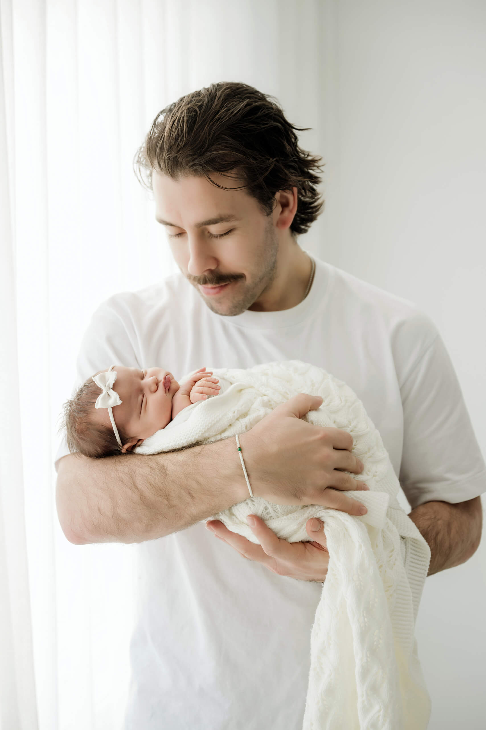 Parent looks down while cradling a newborn in a white knit blanket and bow headband.