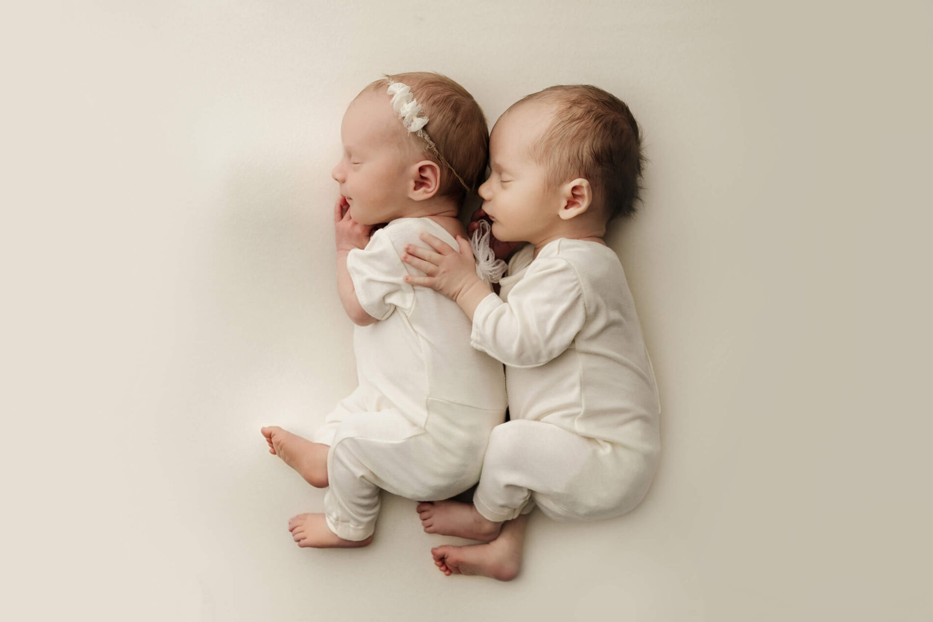 Two newborns with eyes closed lie together in cream outfits, one resting a hand on the other.