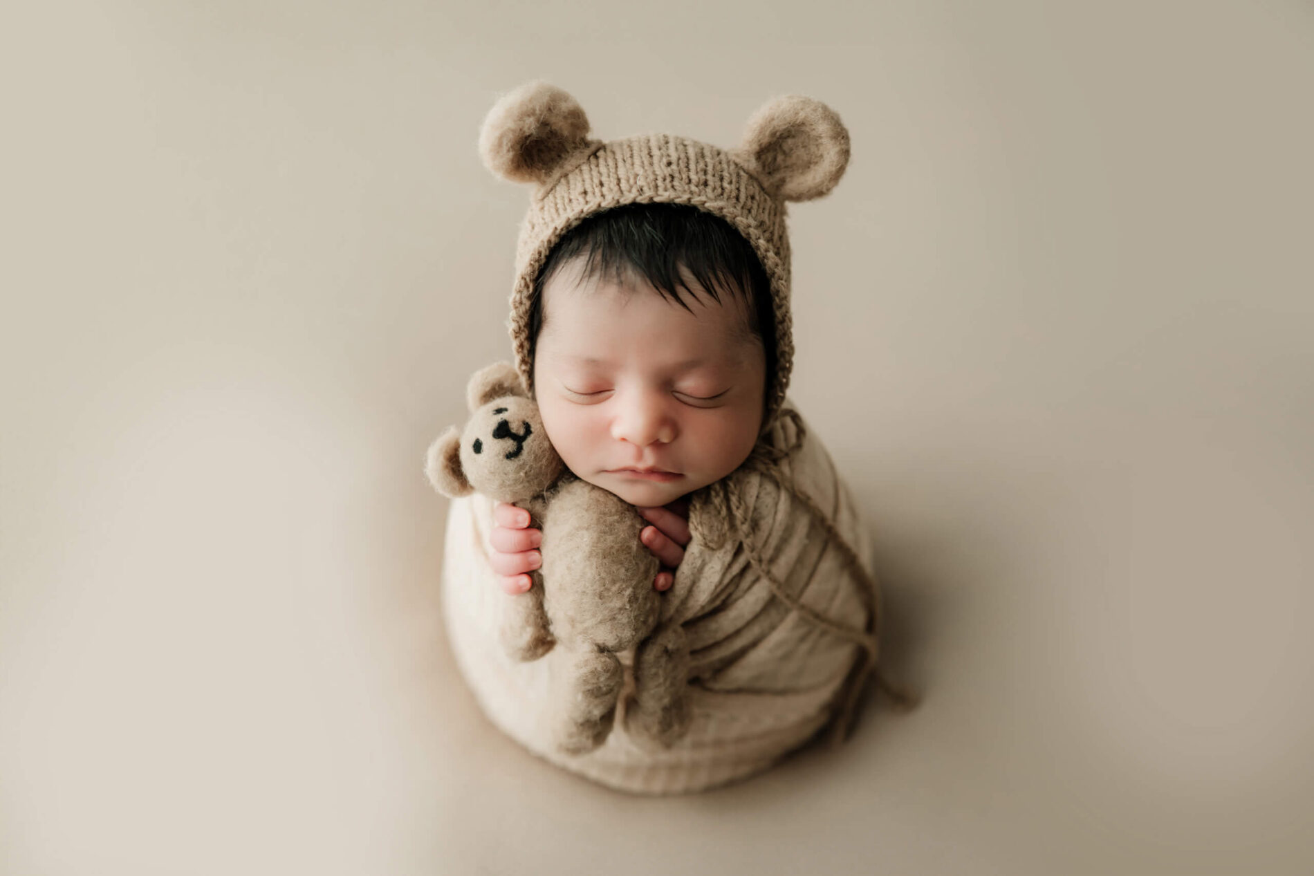 Beige-wrapped newborn with eyes closed wears a bear bonnet and holds a small stuffed bear.