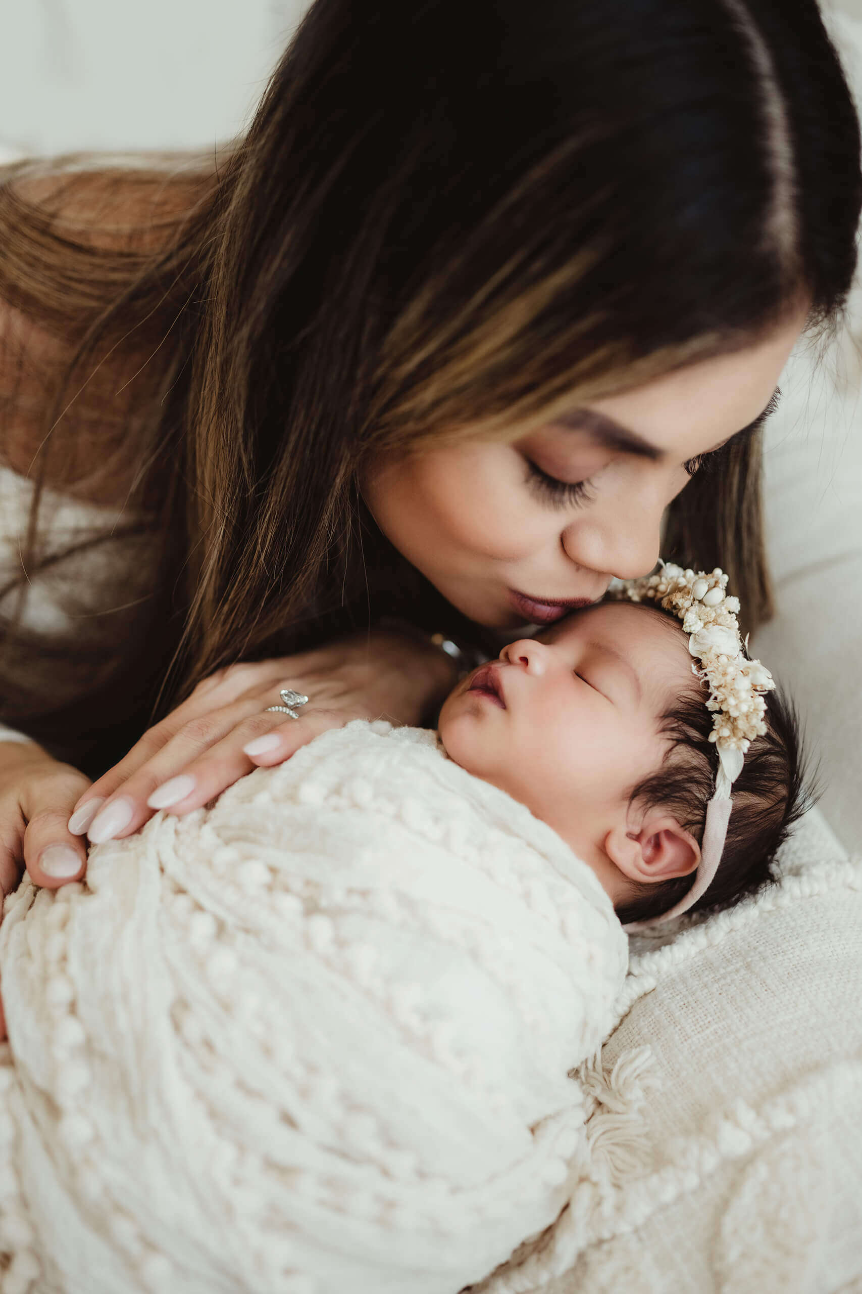 Parent kisses the forehead of a newborn wrapped in white and wearing a cream floral headband.