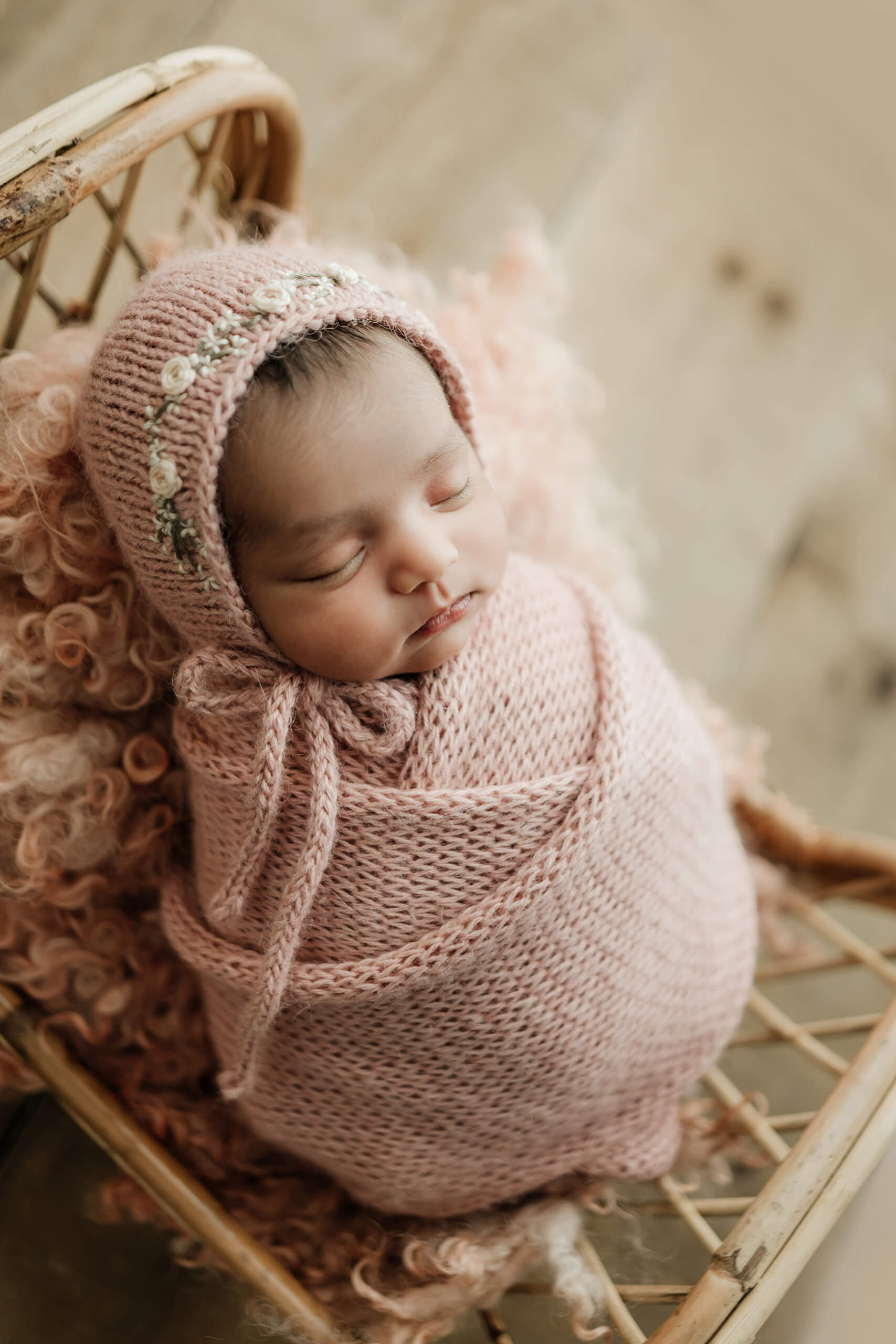 Newborn wrapped in blush pink knit wears a matching floral bonnet while resting in a small rattan bed.