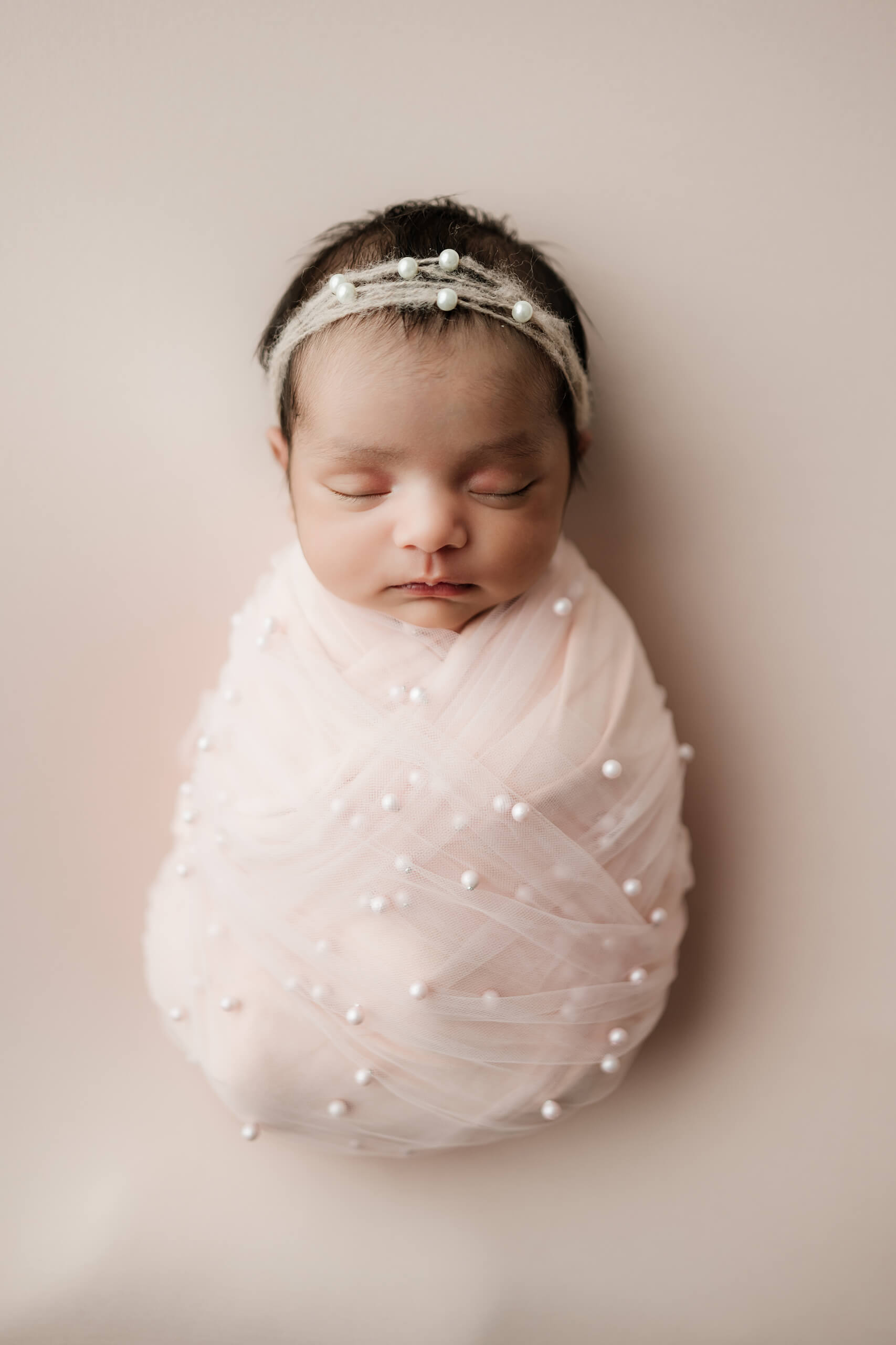 Newborn wrapped in pale pink fabric with pearl details wears a delicate pearl headband.