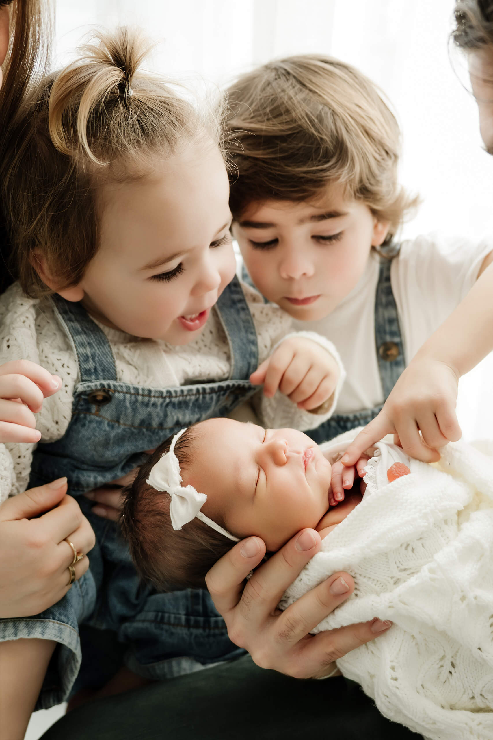 Two young siblings lean close to a newborn wearing a white bow and bundled in a white knit blanket.