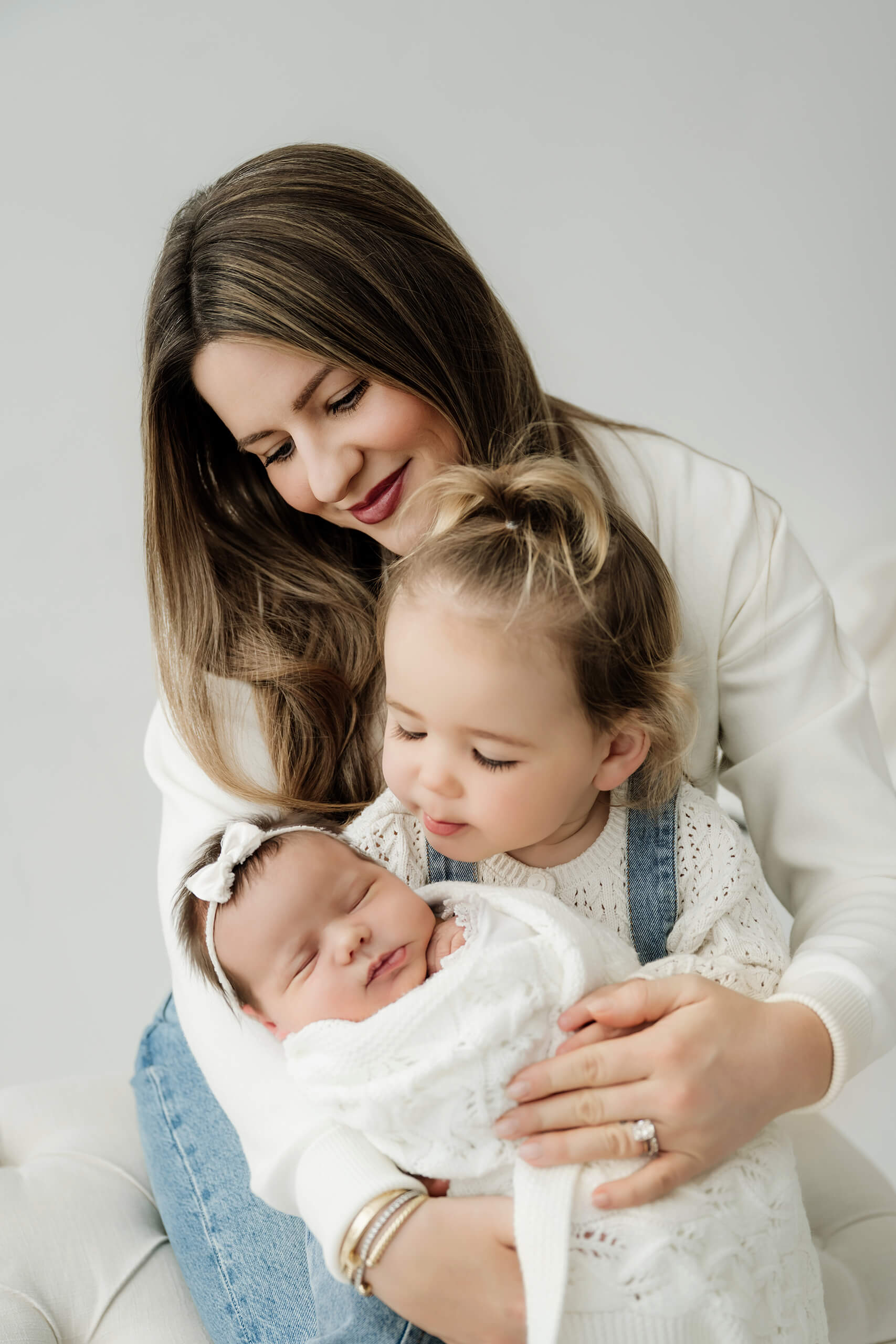Parent holds a sleeping newborn while a young sibling leans close in a bright white studio.