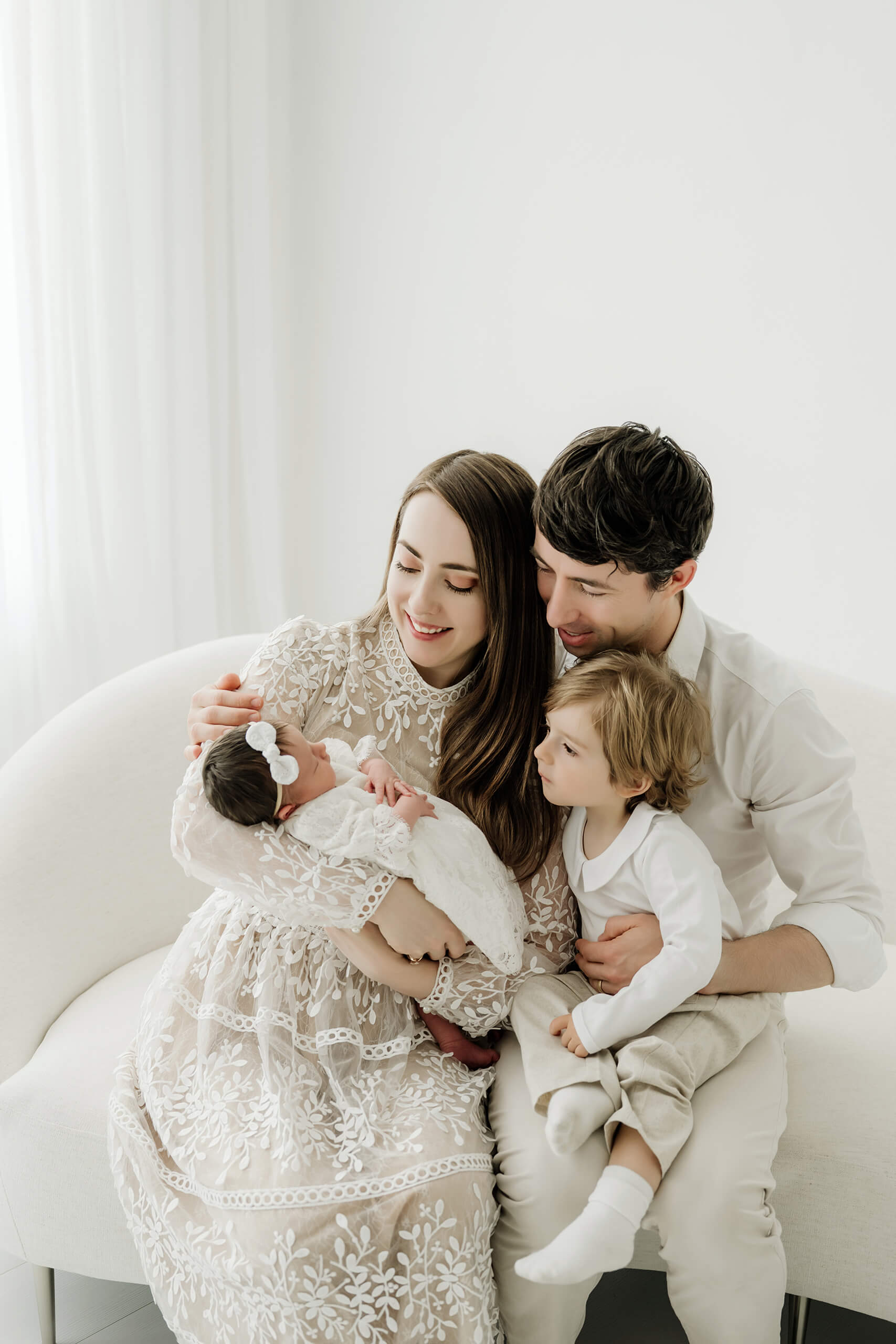 Parents and a young sibling gather around a newborn while seated together on a white studio couch.