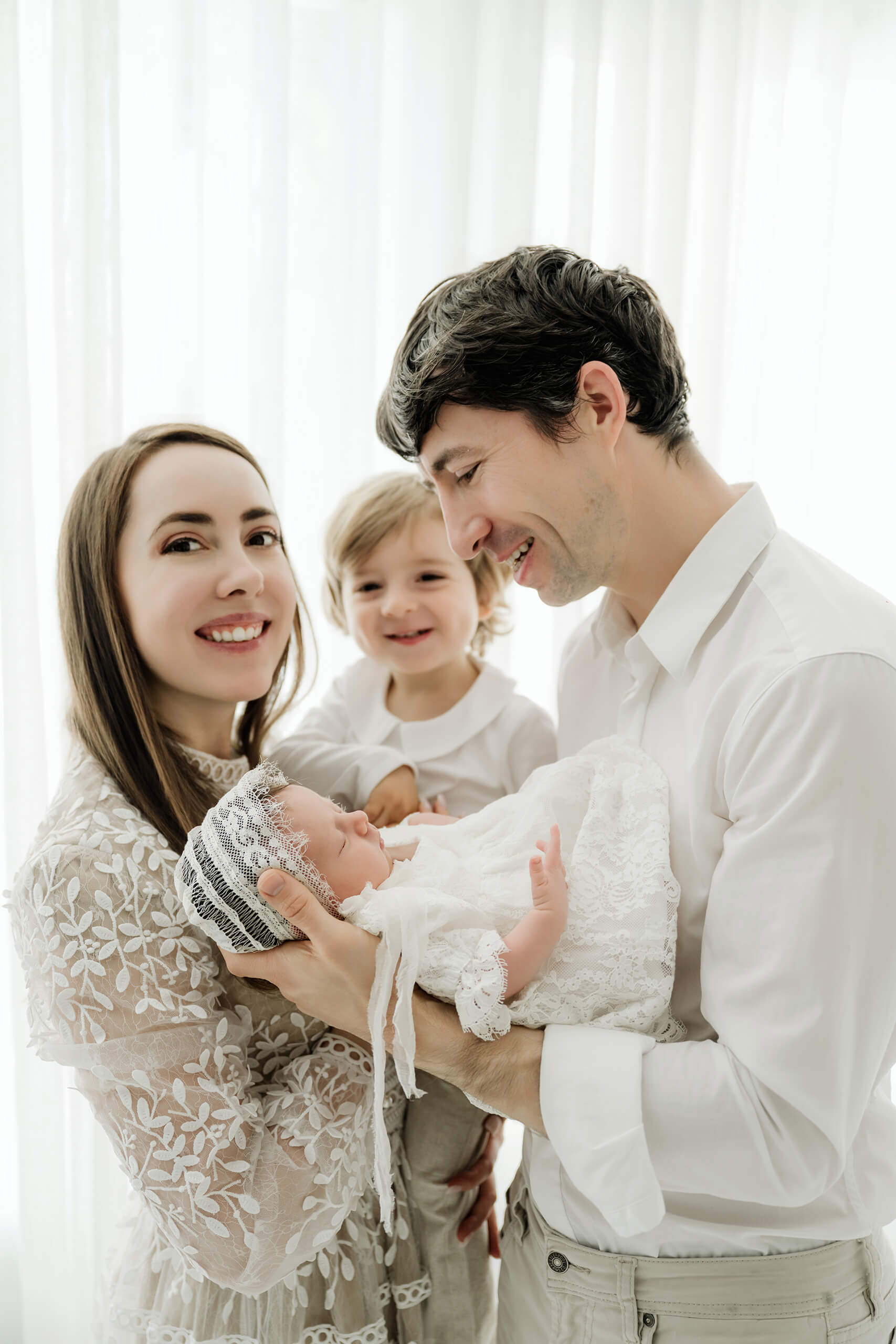 Family of four stands together by a bright studio window as one parent cradles the newborn.