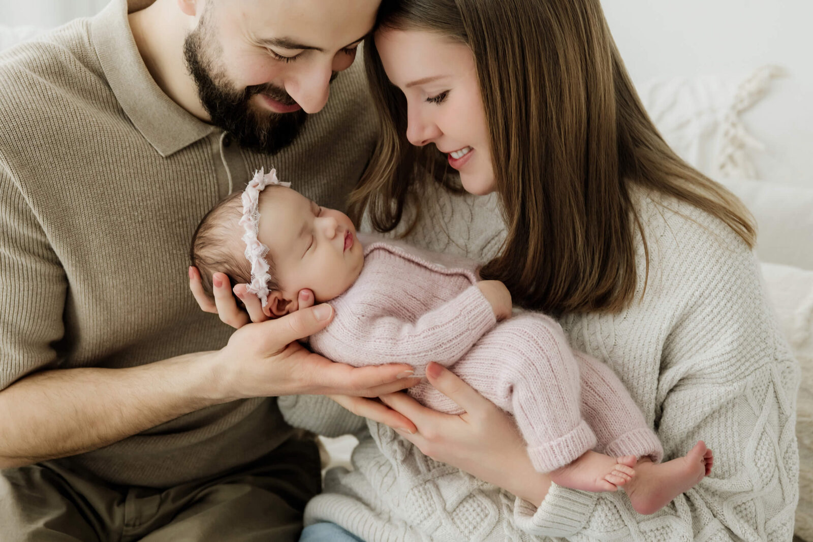 Two parents look down at their newborn in a pale pink knit outfit and floral headband.