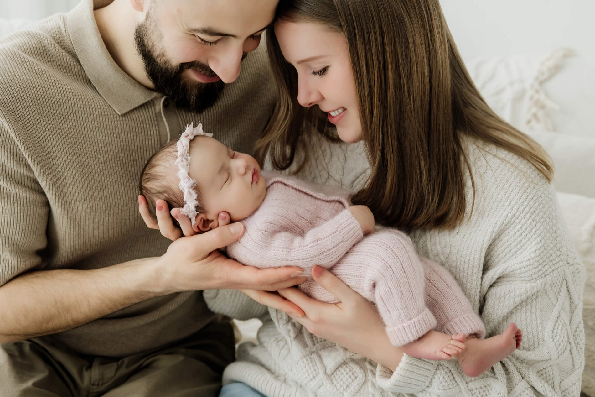 Two parents look down at their newborn in a pale pink knit outfit and floral headband.
