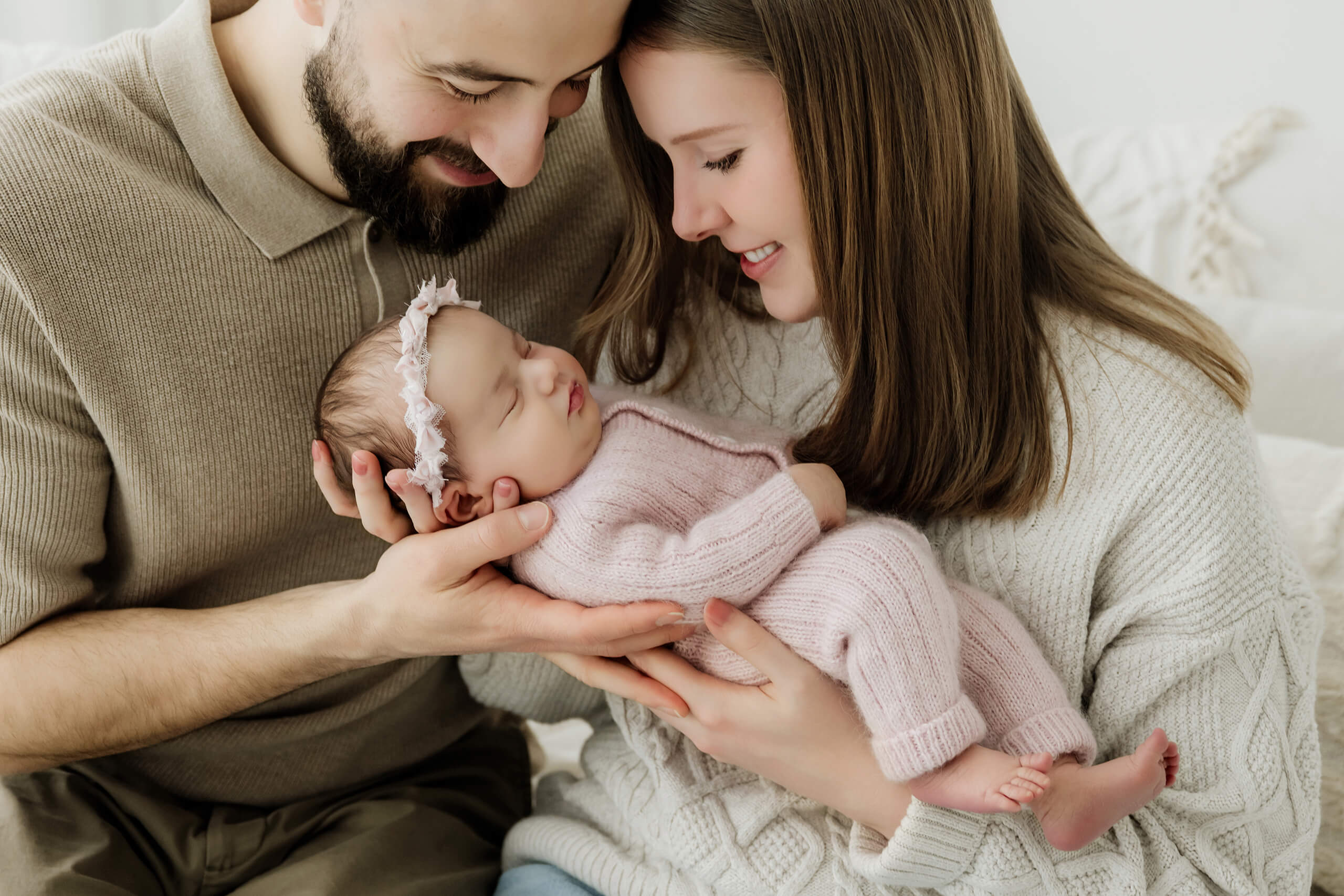 Two parents look down at their newborn in a pale pink knit outfit and floral headband.