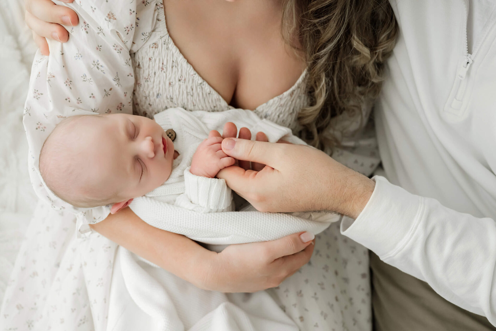Newborn with eyes closed is cradled in one parent’s arms while another holds the baby’s hand.
