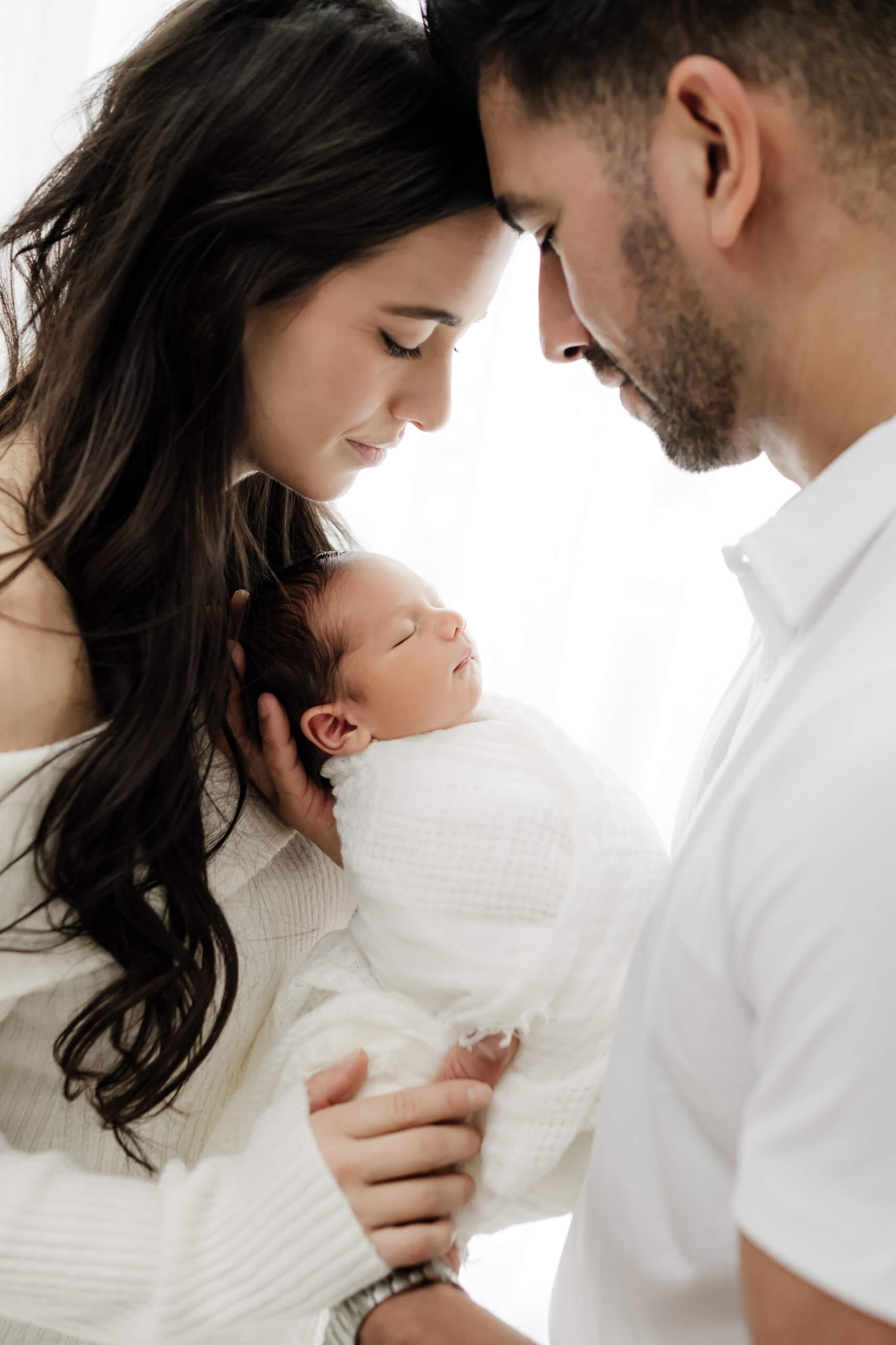 Two parents lean close over their newborn wrapped in white against a bright studio background.