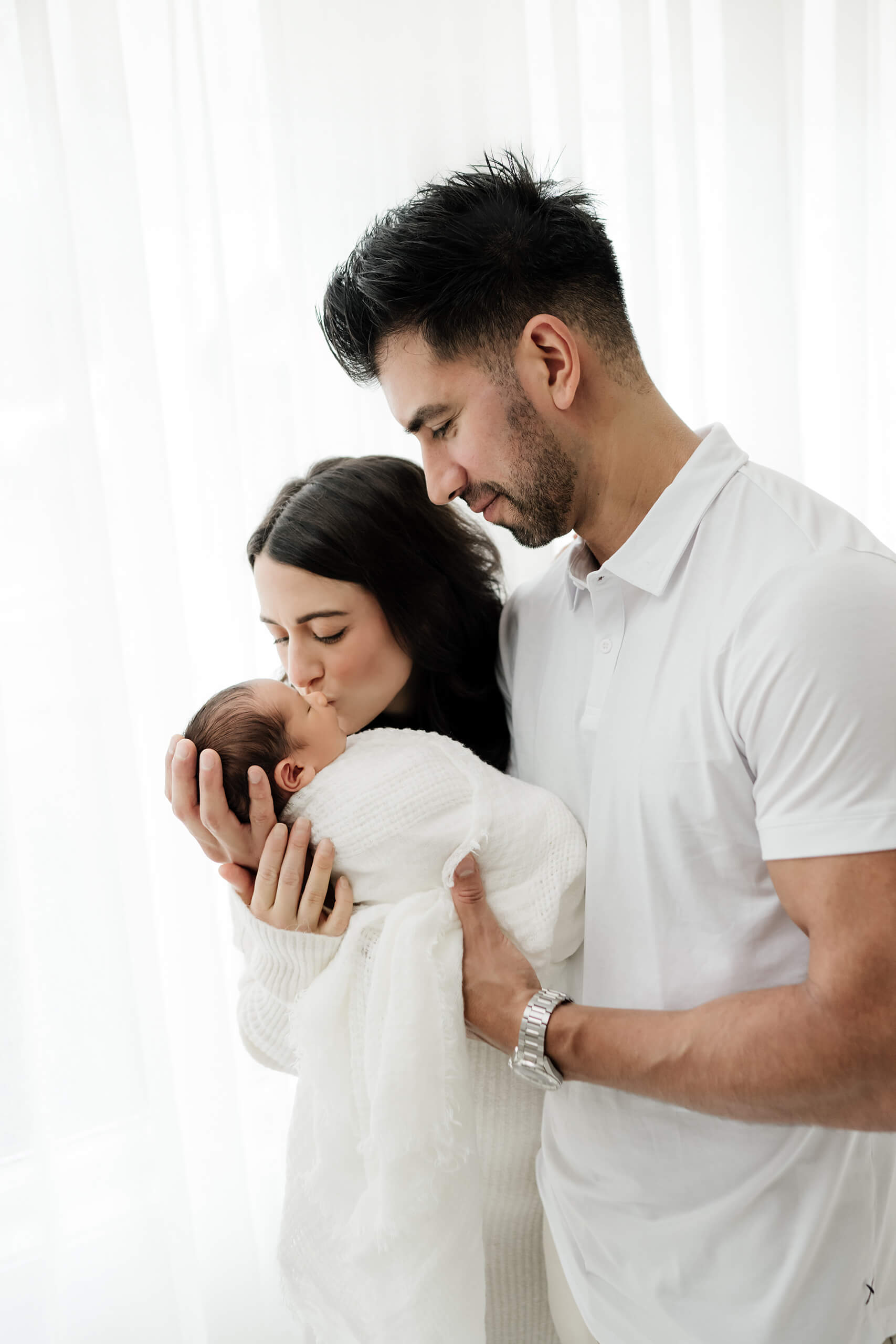 One parent kisses a white-wrapped newborn while the other supports the baby beside them.