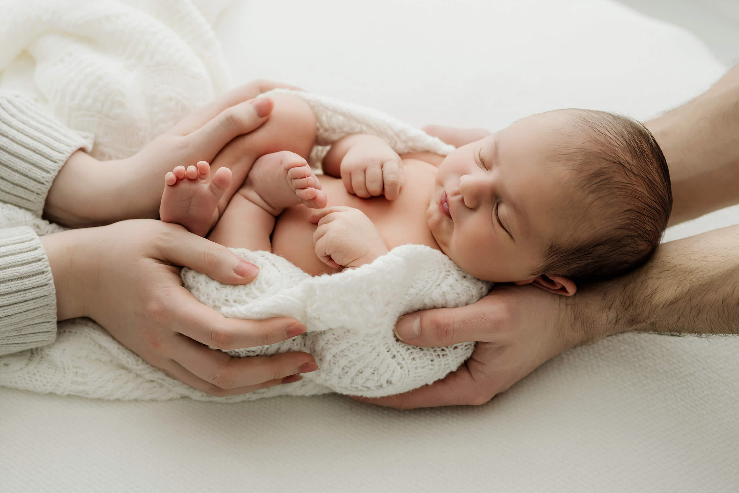 Newborn with eyes closed rests across both parents’ hands, with bare feet and curled fingers visible.