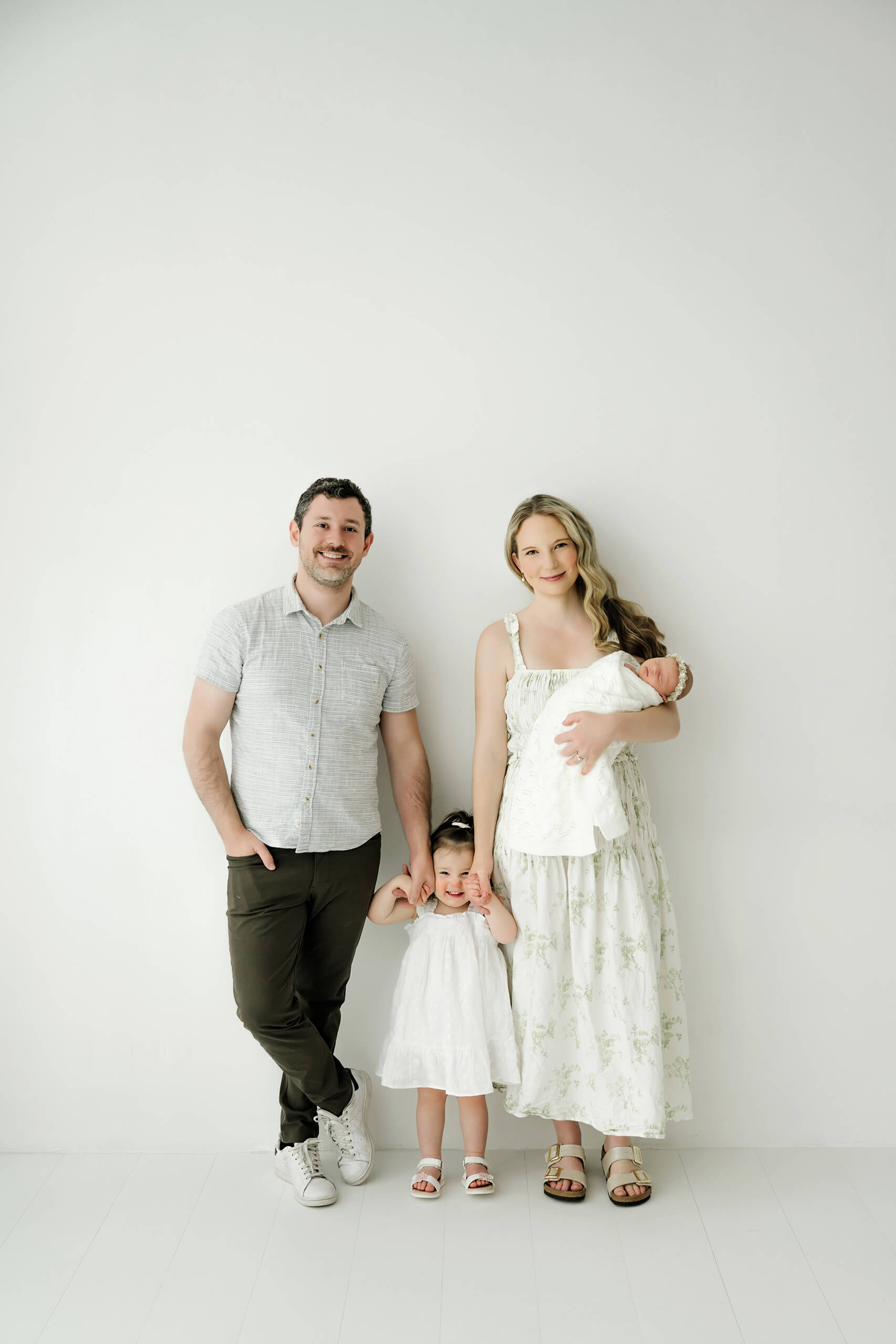 Family of four stands against a white wall while one parent holds the newborn.