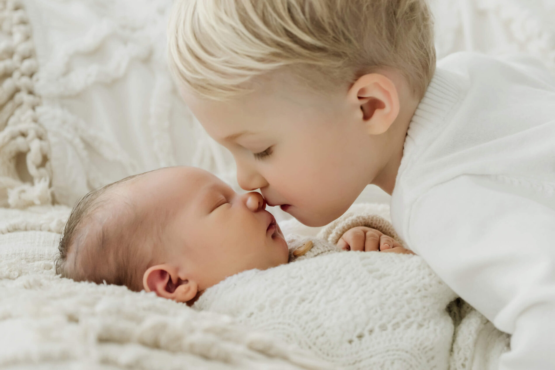 Young sibling leans nose-to-nose with a newborn supported in an adult’s arms.
