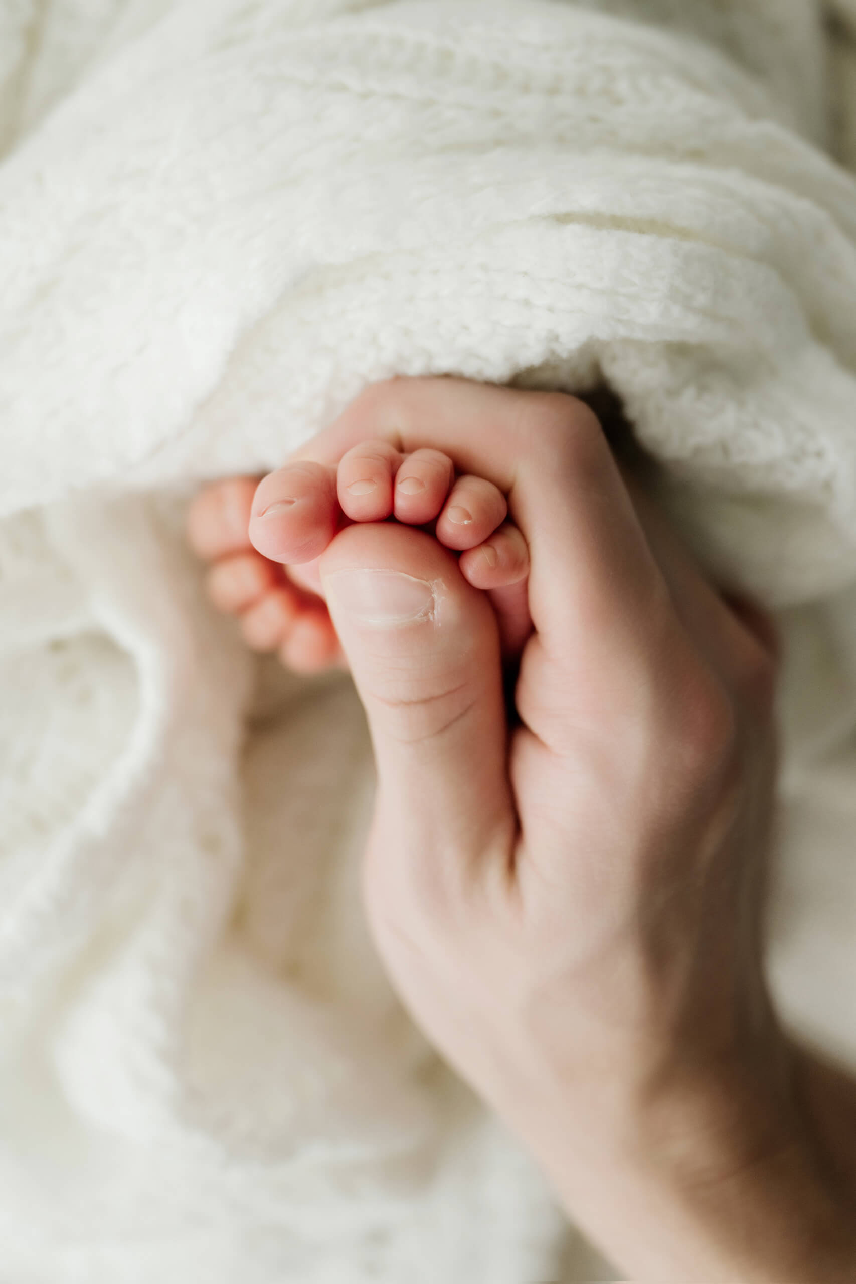 Close-up of an adult hand holding a newborn’s tiny toes against a white knit blanket.