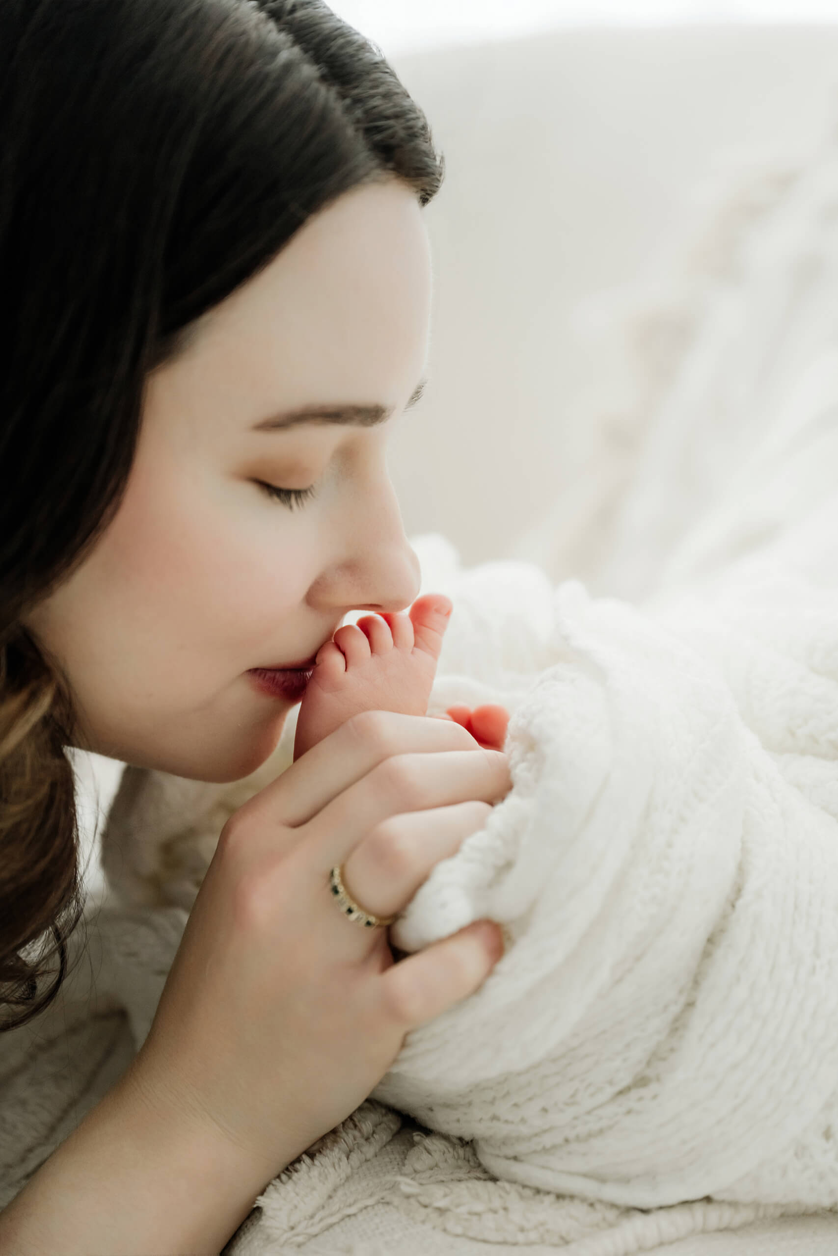 Parent kisses a newborn’s toes while holding the baby beneath a white knit blanket.