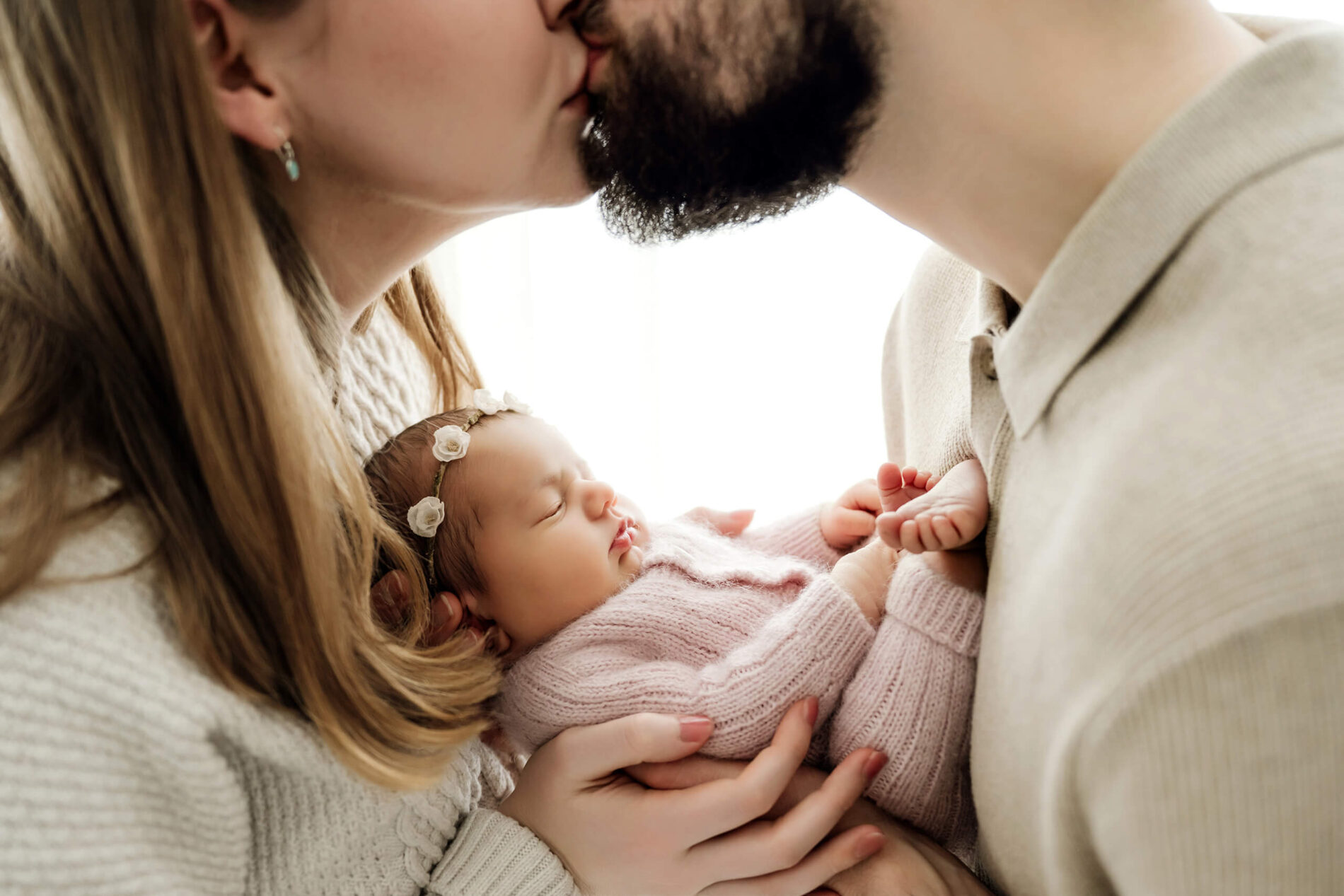 Two parents kiss above their newborn, held between them in a pale pink knit outfit and floral headband.