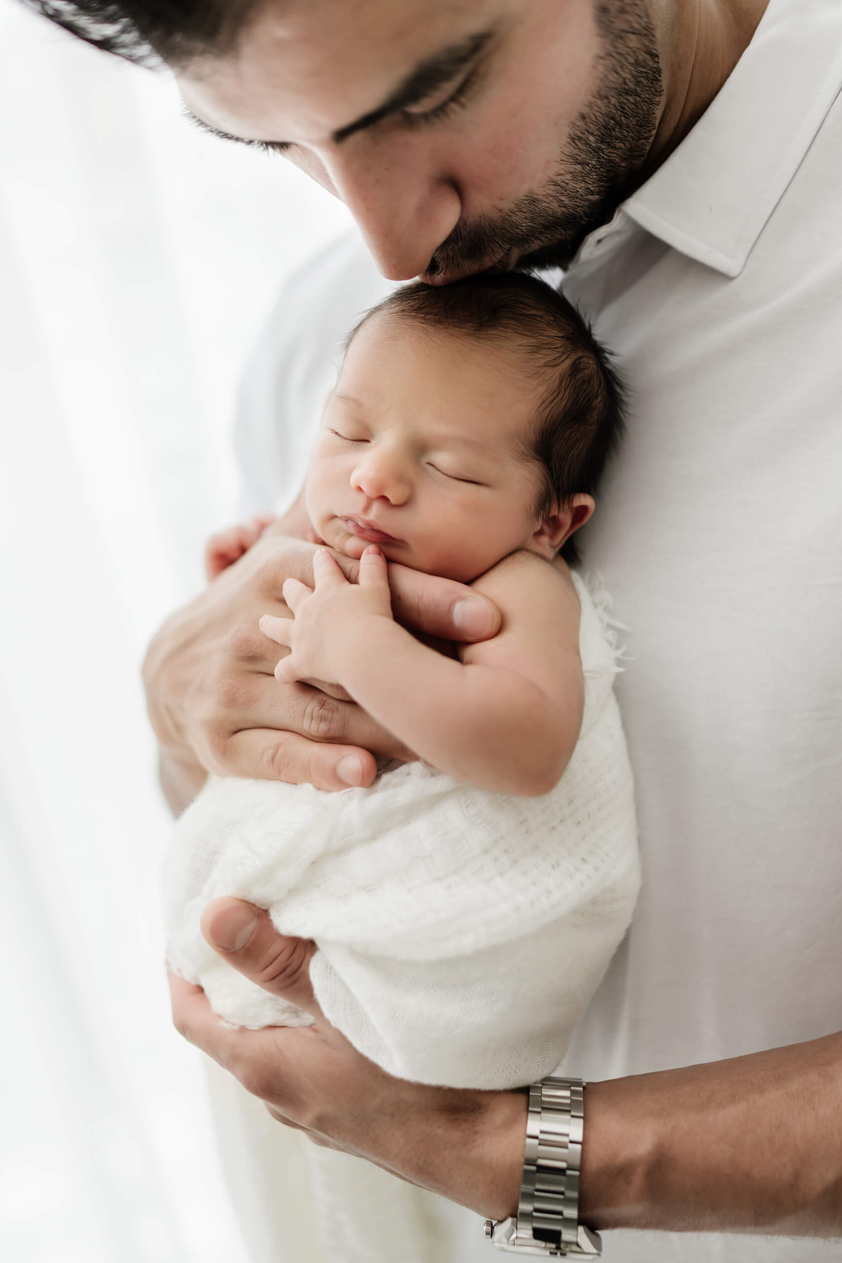 Parent holds a newborn with eyes closed against their chest and kisses the baby’s head.