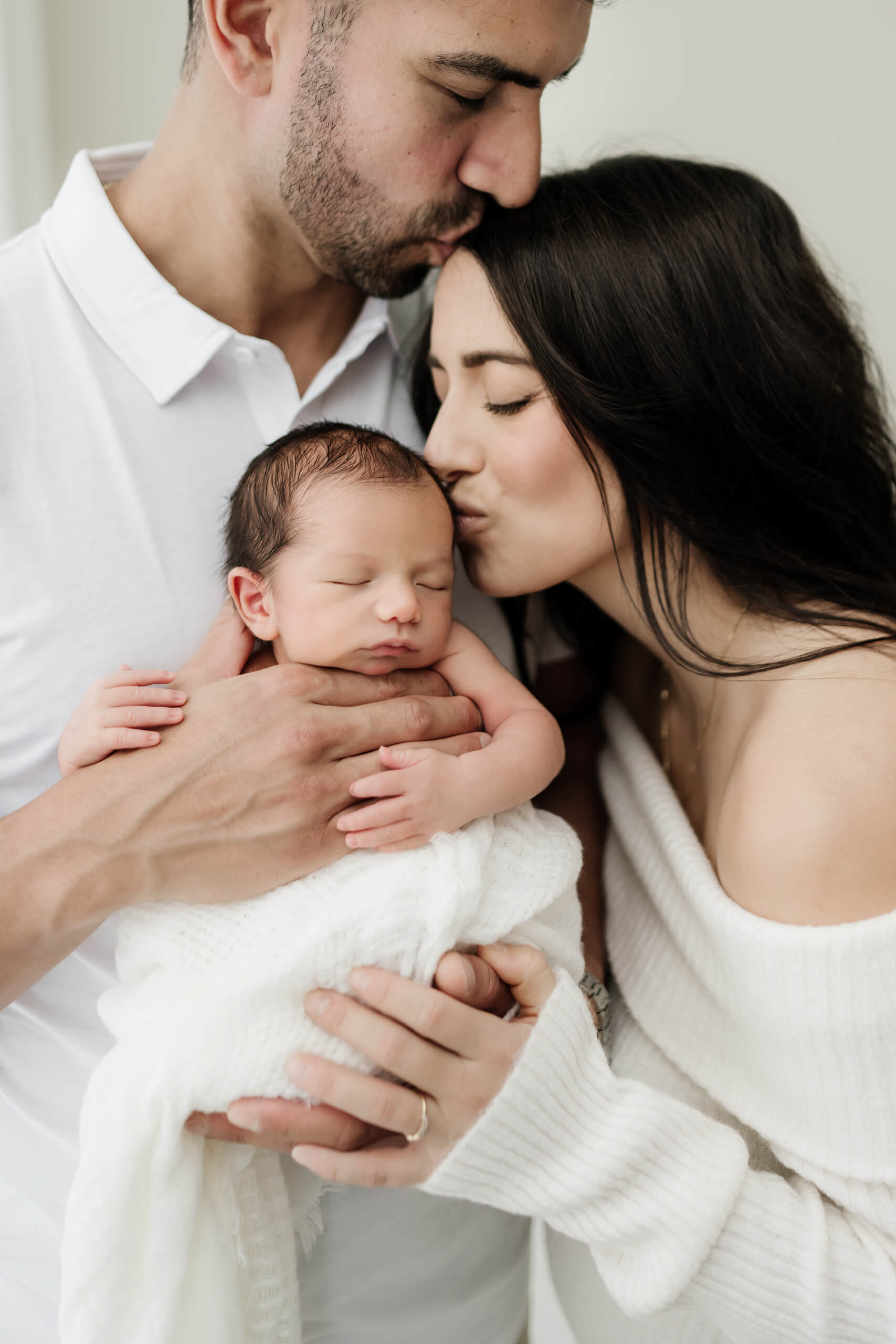 Two parents hold their newborn while one kisses the baby and the other kisses their partner’s forehead.